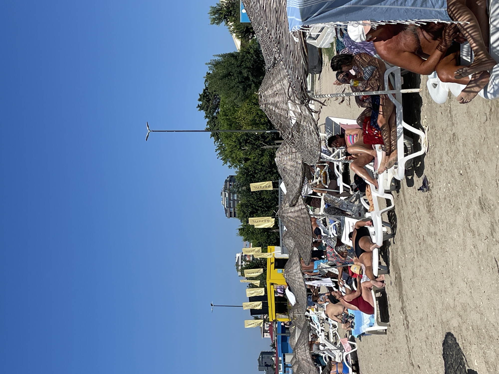 The image depicts a crowded beach scene on a sunny day in Kadıköy, Turkey. Numerous beachgoers are seen relaxing on lounge chairs under thatched umbrellas. People are dressed in swimwear, and many are sunbathing or engaging in conversations. A bright yellow structure with yellow flags bearing text is visible in the background, suggesting a vendor or service area. Buildings and trees are also present in the distance. The sand on the beach is light brown, and the sky is clear blue. The composition suggests a typical summer day at a popular beach resort.