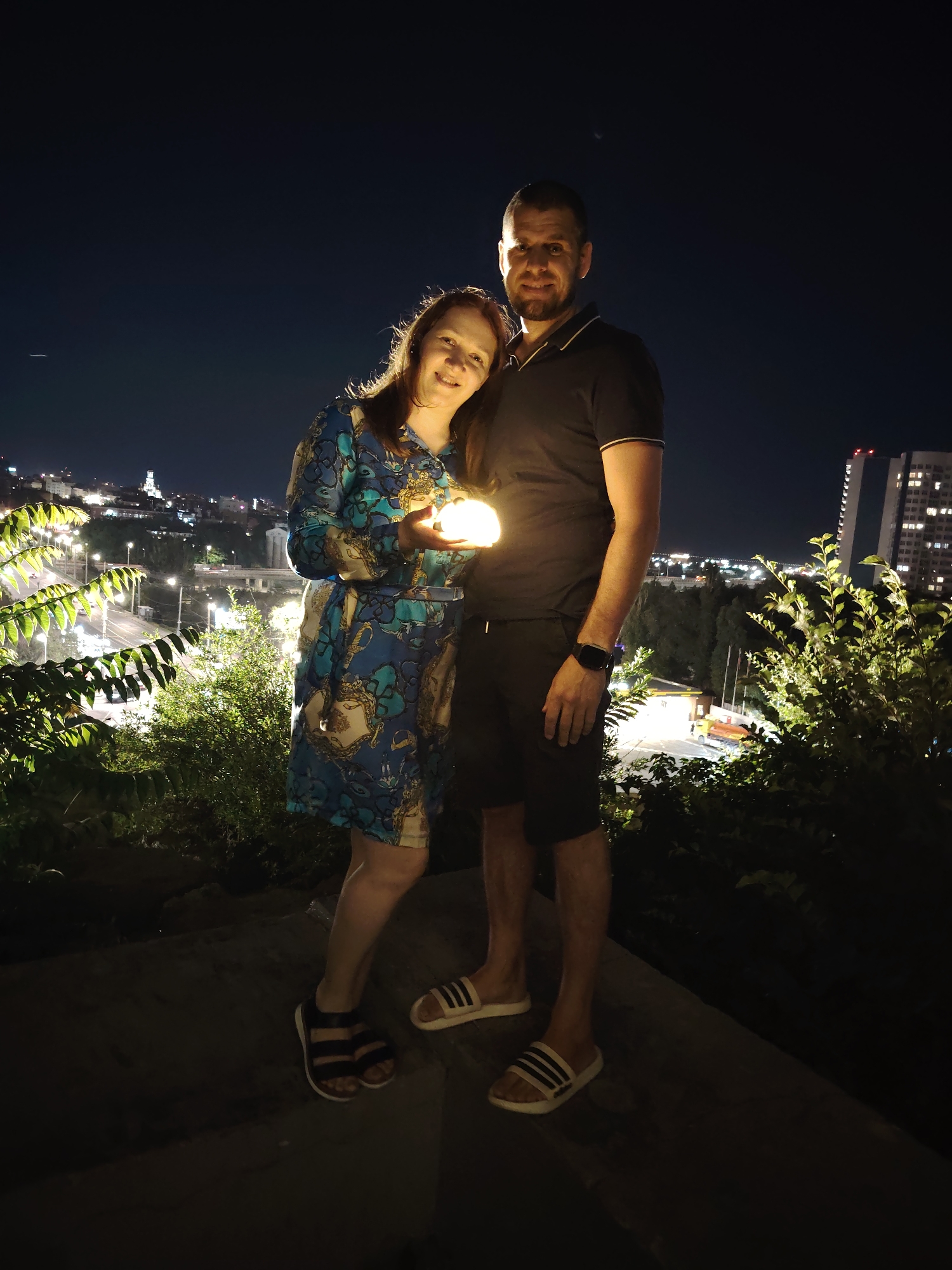 A man and a woman are standing on a concrete surface at night with a city skyline in the background. The woman is wearing a blue and gold patterned dress and holding a glowing orb. The man is wearing a black polo shirt and black shorts, with a smartwatch on his left wrist. They are both looking at the camera and smiling. In the background, there are buildings with illuminated windows, streetlights, and lush green trees and bushes in the foreground. The city appears to be hilly, with a bridge visible in the distance. This appears to be a nighttime photograph taken from an elevated position overlooking the city of Rostov-on-Don, Russia, likely from a viewpoint like the one on the embankment near the St. Nicholas Church or a similar vantage point.