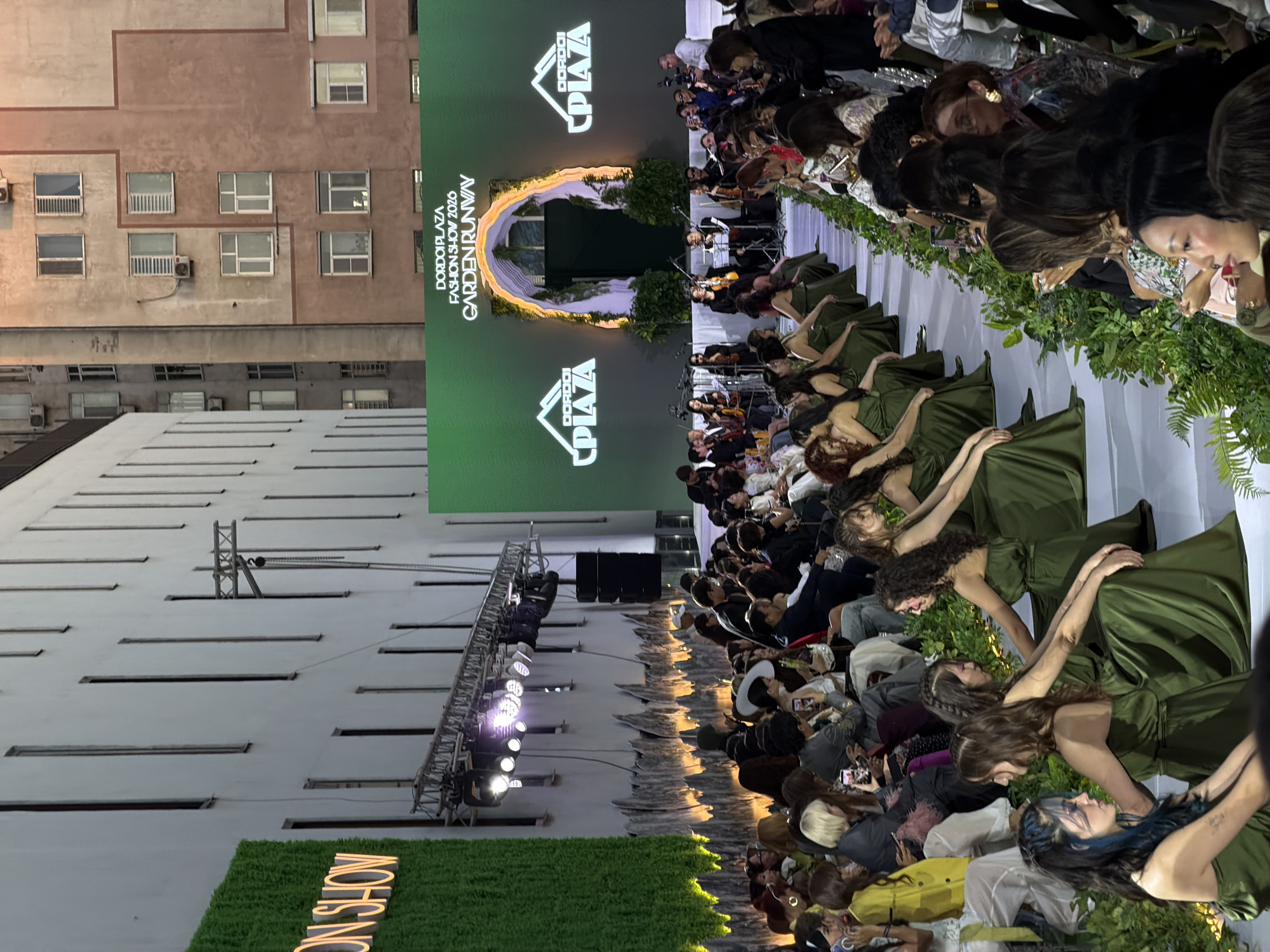 The image shows a fashion show, possibly at night, identified by the words "GARDEN RUNWAY" on a turf-covered display. The location appears to be Bishkek City, Kyrgyzstan, as indicated by "DORDOI PLAZA" on a large screen behind the runway. The runway is lined with seated audience members and greenery. Models in long, olive-green dresses are visible on the runway. A string orchestra performs on the side of the runway. Stage lights and rigging are present, and a building with windows is in the background. The entire scene suggests an outdoor fashion event in an urban environment.