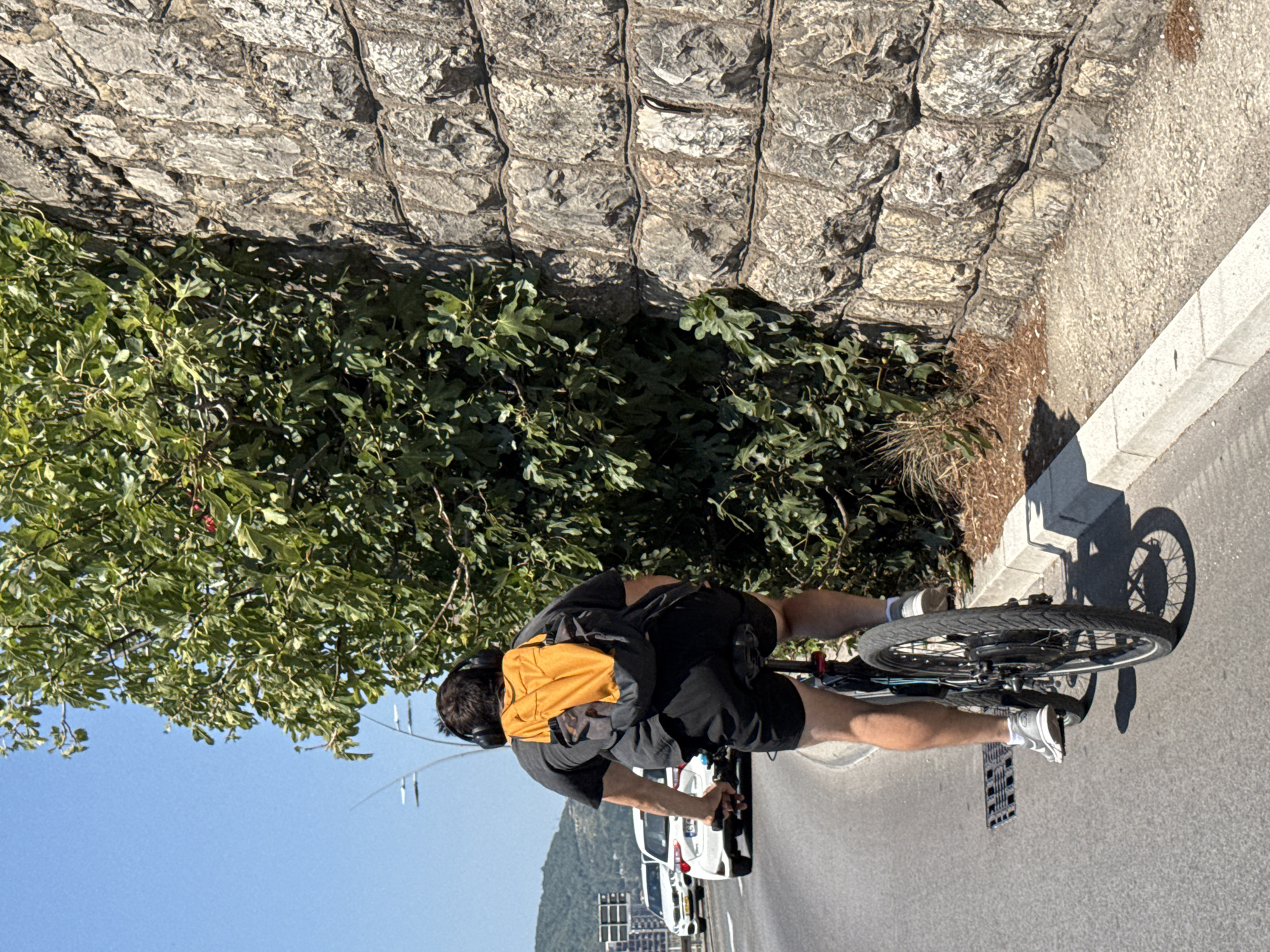 A man wearing headphones and an orange backpack rides a bicycle on a paved road next to a stone wall and dense foliage. He is wearing a dark t-shirt and black shorts. The road has a grated drain cover on the left side. In the background, white cars are driving on the road. The sky is clear and blue. The scene appears to be on a street in Budva, Montenegro, likely along a coastal road given the stone wall and vegetation.
