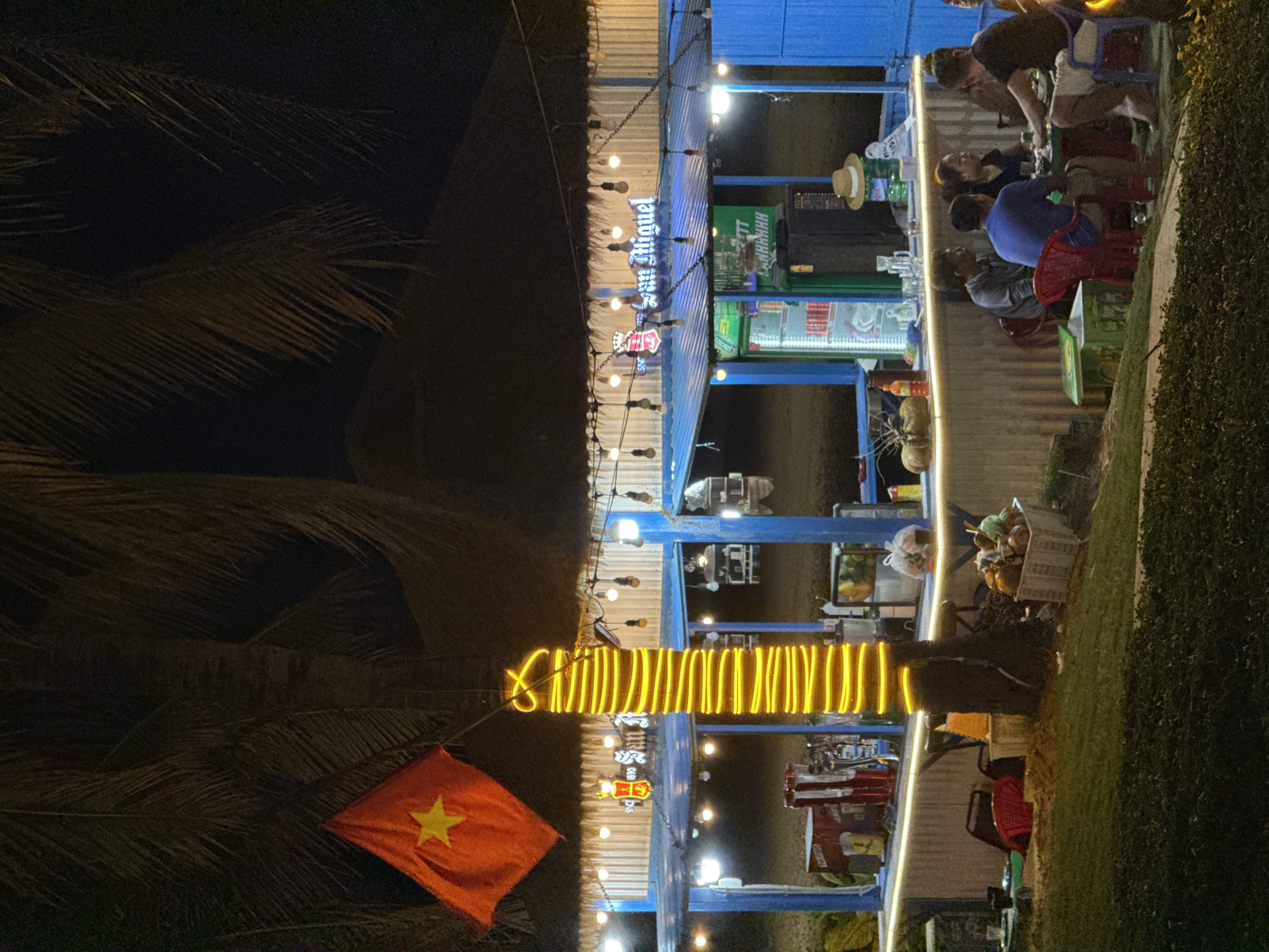 The image captures a lively outdoor night scene at what appears to be a series of food or beverage stalls. Multiple open-air stalls with blue structural elements and corrugated metal siding are visible on the right, brightly illuminated by string lights with exposed bulbs and internal lighting from coolers and counters.
Several people are present, socializing and engaging in activities typical of an outdoor bar or eatery. Some individuals are seated at tables on red plastic chairs, while others stand, possibly serving or interacting. A person in a blue shirt is seated with their back to the viewer, and another individual wearing a dark shirt and a light-colored apron stands near a counter, seemingly attending to the establishment. A straw hat rests on a counter in one of the stalls.
To the left, a large palm tree is prominent, its fronds silhouetted against the dark night sky. The tree's trunk is decoratively wrapped with bright yellow string lights. Below the palm fronds and to the left of the illuminated stalls, a red flag with a large yellow star, clearly identifying it as the national flag of Vietnam, hangs.
The ground area is lined with green shrubbery. Baskets filled with fruits, including coconuts, are visible near the stalls, along with various bar equipment. A prominent sign above one stall displays the text "San Miguel." Another green sign features partially visible text that reads "HNNH HHH." The scene conveys a casual and relaxed atmosphere, characteristic of an evening gathering in a tropical coastal setting like Đà Nẵng, Vietnam.
