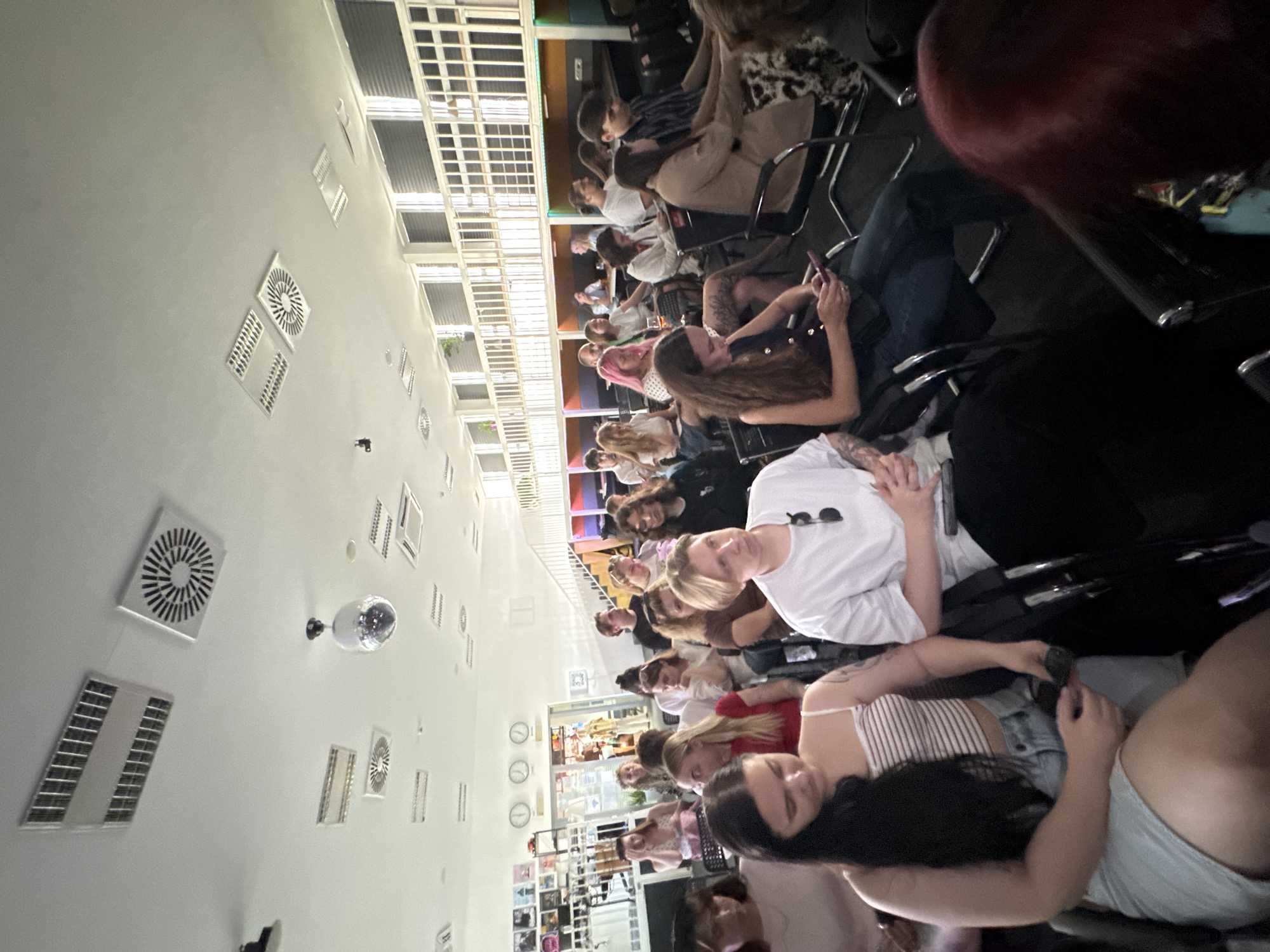 A medium, high-angle shot displays a group of people seated indoors, seemingly an audience in an event. The setting appears to be a modern space, potentially a venue or lounge in Bar, Montenegro. A white ceiling with circular ventilation grates and a disco ball indicates an entertainment or gathering space. Attendees, predominantly women, are seated in chairs. The interaction includes some on their mobile phones, others talking, and some watching the presentation. The people seem to be interacting with each other.