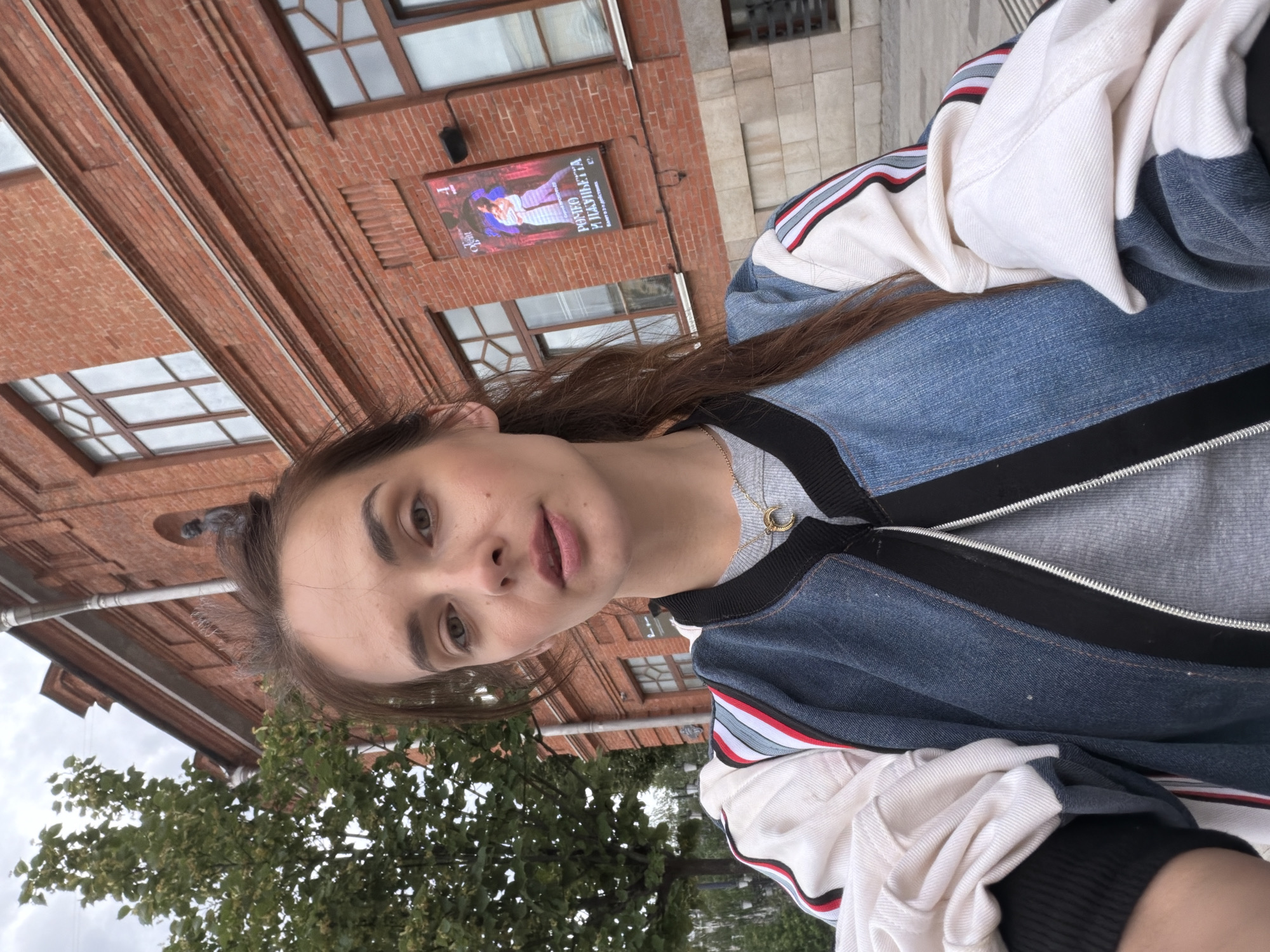 A young woman with brown hair pulled back in a ponytail takes a selfie outdoors in front of a red brick building. She is wearing a grey t-shirt and a denim and white bomber jacket with red, white, and blue striped detailing on the sleeves. A silver necklace with a crescent moon pendant is visible around her neck. A poster is affixed to the brick building behind her, depicting two figures in a theatrical pose, with Russian text that translates to "Romeo and Julietta" and "Ballet in two acts." The background also shows a tree with green foliage and a cloudy sky. This scene is located in Ufa, Russia, specifically near the Ufa State Tatar Theater "Nur."