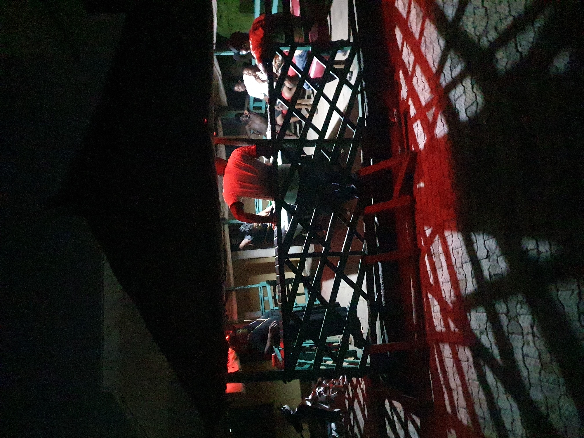 The image shows a multi-level wooden structure with people on different levels. The structure is surrounded by darkness, with some areas illuminated by red light. Shadows of the structure are cast on a nearby wall. People are wearing various colors of clothing, including red, white, and dark colors. The location appears to be Lokoja, Nigeria.