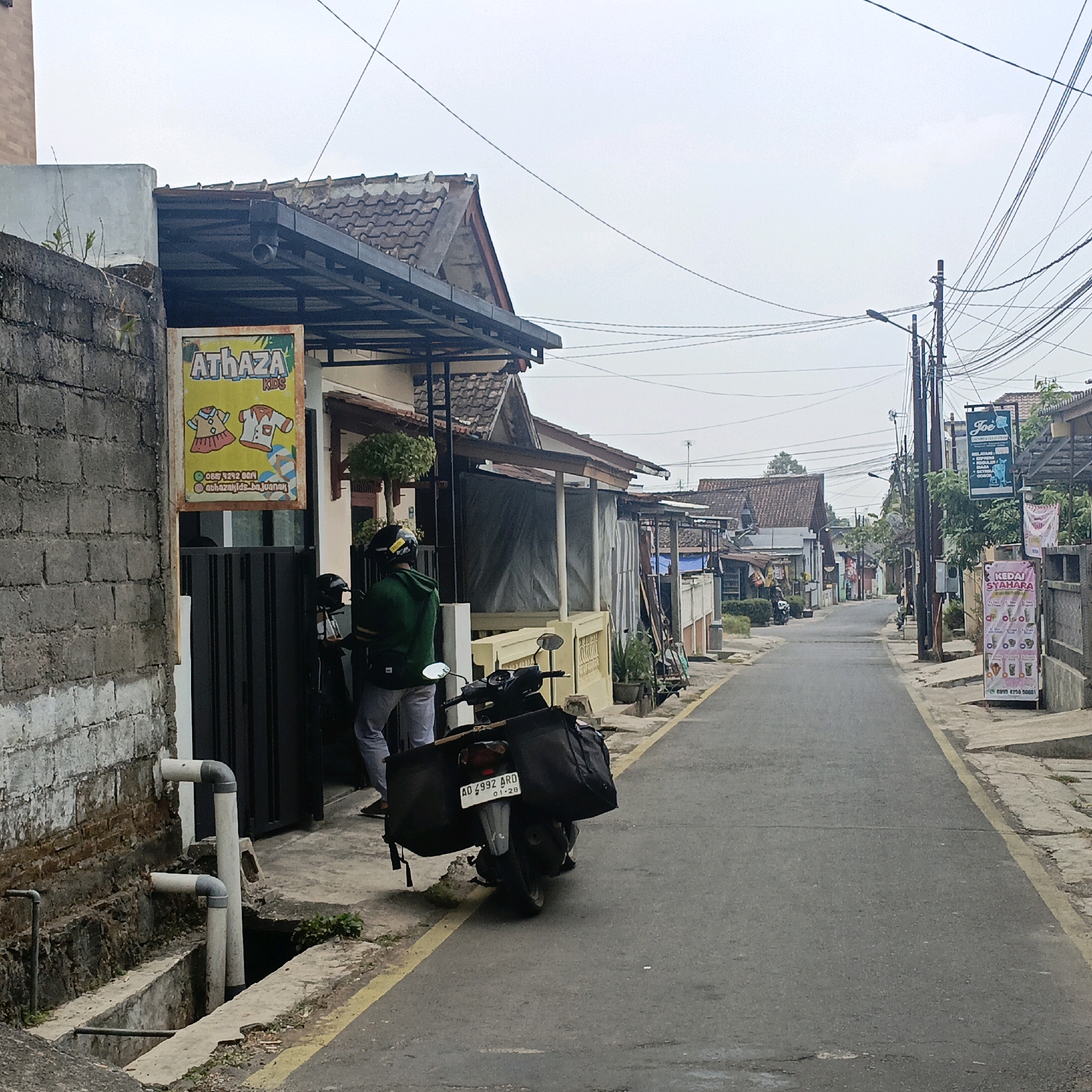 The image depicts a narrow street in Salatiga, Indonesia, likely a residential or commercial area with a mix of single-story buildings. A person wearing a green hoodie and a helmet is standing next to a parked motorcycle, which has a large black bag attached to the rear. A sign for "Athaza Kids" is visible on the left side of the street, featuring cartoon drawings of children's clothing. On the right side of the street, a sign advertises "Kedai Syahara" with images of various drinks. Utility poles with a tangle of wires dominate the sky. The pavement is asphalt with a yellow line marking the edge of the road. Several buildings line the street, featuring tiled roofs and various architectural styles. The overall scene suggests a typical day in this Indonesian city.