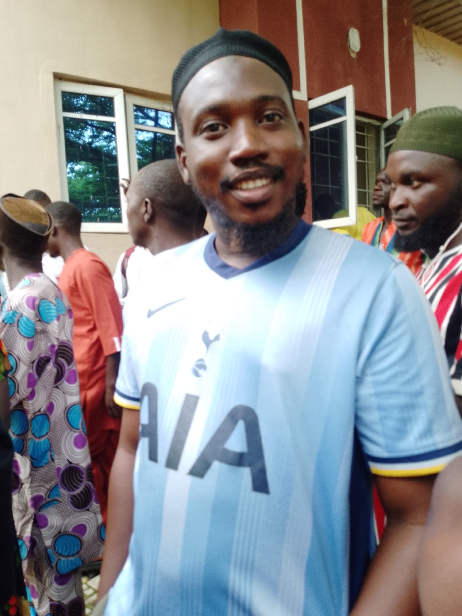 A group of people are gathered outdoors in Akwanga, Nigeria. The subject in the foreground is a man wearing a light blue t-shirt with a white Nike logo and the word "AIA" prominently displayed. He is also wearing a black skullcap and has a beard. He is smiling and looking directly at the camera. Behind him, other individuals are visible, some wearing traditional African clothing, including a woman in a colorful patterned dress. Others are wearing shirts and caps, with one man to the right wearing a green cap. The background shows a building with windows and a light-colored wall. The scene appears to be during daylight hours. The overall mood seems casual and social, with the man in the foreground appearing happy.