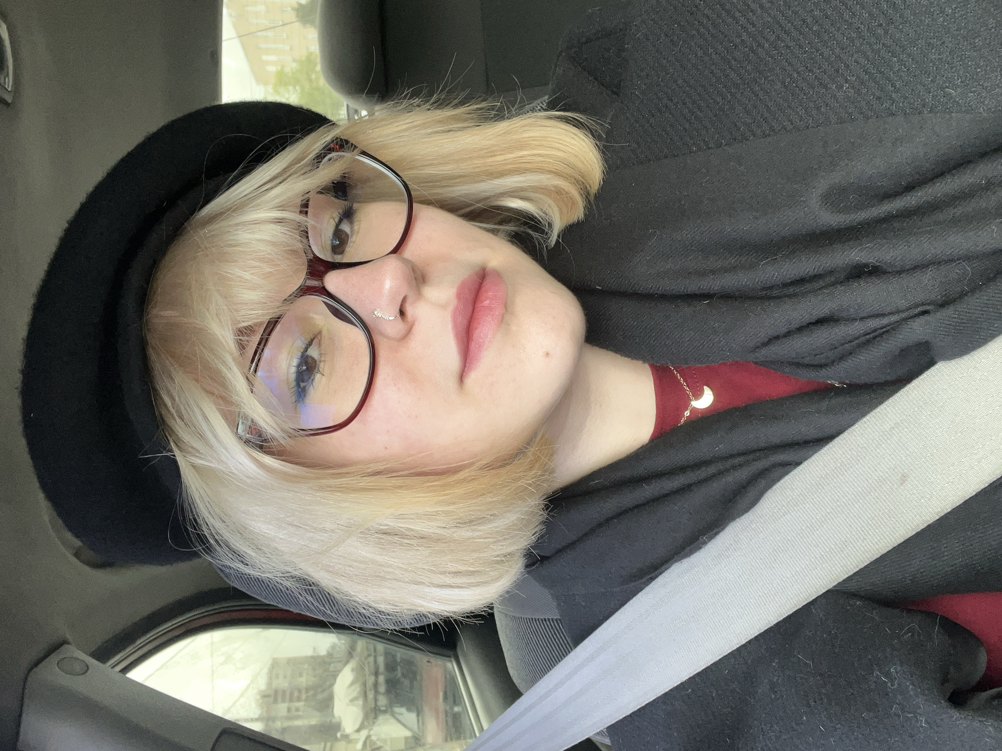 A young woman is captured in a medium close-up selfie, seated inside a car. She appears to be in her late teens or early twenties, featuring short, light blonde hair with bangs and wearing large glasses with dark red frames. A small, silver nose ring adorns her right nostril, and her lips are subtly colored with a soft pink hue. Her expression is neutral and calm, as she gazes directly towards the camera.
She is dressed in a dark red top, with a black beret on her head and a black outer garment, possibly a coat or a thick scarf, draped around her shoulders. A delicate gold-colored necklace with a crescent moon pendant is visible on her neck. A light grey seatbelt runs diagonally across her chest and shoulder, indicating her position as a passenger in the vehicle.
The background reveals the car's interior, including parts of the dark fabric seat and interior trim. Through the car window, blurred shapes of buildings and an urban environment suggest it is daytime. The lighting is soft and diffused, implying an overcast day. There is no visible text within the image.