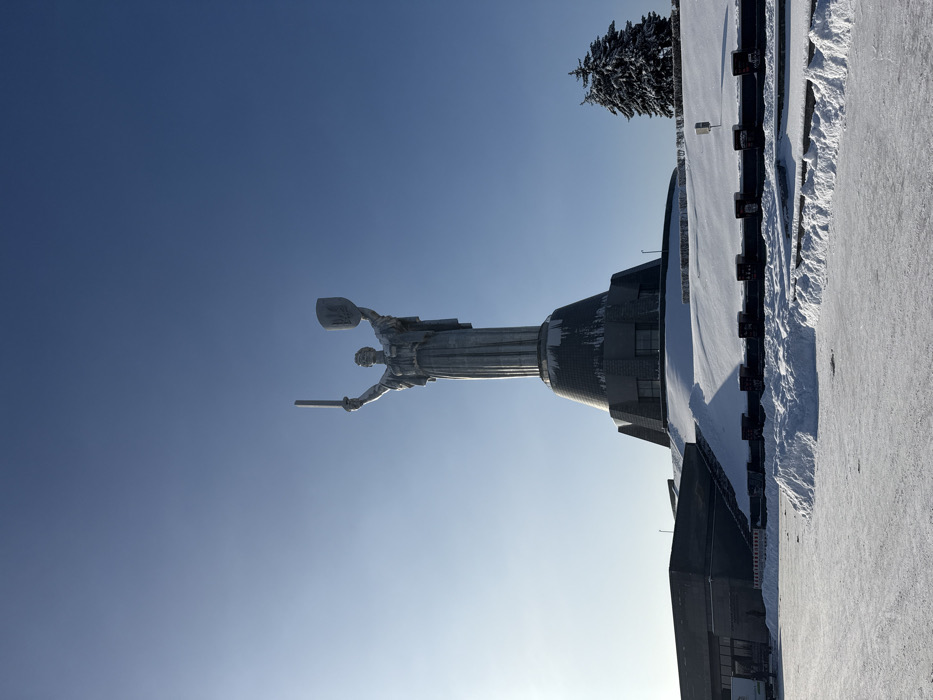 The image displays the Motherland Monument (Rodina Mat) located on the grounds of the National Museum of the History of Ukraine in the Second World War, in Kyiv, Ukraine. The scene is a winter landscape under a clear, bright blue sky.
The central subject is a colossal metallic statue of a female figure, depicted mid-stride, holding a sword vertically aloft in her raised right hand and a shield bearing the Ukrainian trident emblem in her raised left hand. The statue stands on a multi-tiered, conical pedestal made of dark stone or metal, with some snow and ice accumulation visible on its lower sections.
The surrounding environment is covered in snow. A large, open, snow-dusted ground area occupies the foreground, with distinct piles of cleared snow along its edges. To the immediate right of the monument's base, a dark evergreen tree heavily laden with snow is visible. On the far left, a dark, multi-story building structure with architectural elements is partially visible, also dusted with snow. No people or discernible actions are visible in the frame. Sunlight appears to originate from the left, casting faint shadows.