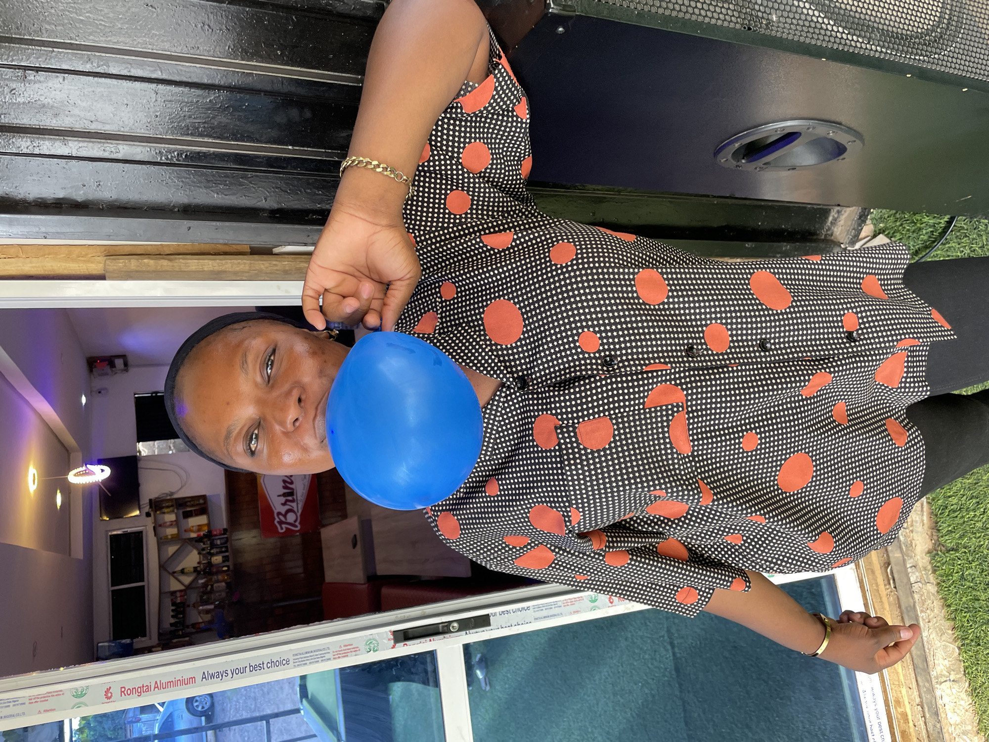 The image depicts a woman holding a blue balloon, seemingly indoors, with a bar visible in the background. She is wearing a black and white patterned shirt with orange circles. A "Brin" sign is partially visible, suggesting the bar's name. A window frame with "Rongtai Aluminium Always your best choice" is visible. This is potentially a bar/restaurant within the city of Jos, Nigeria, given the "IGERIA COLTO" and "NIGERIA COLTO" texts on the window frame.