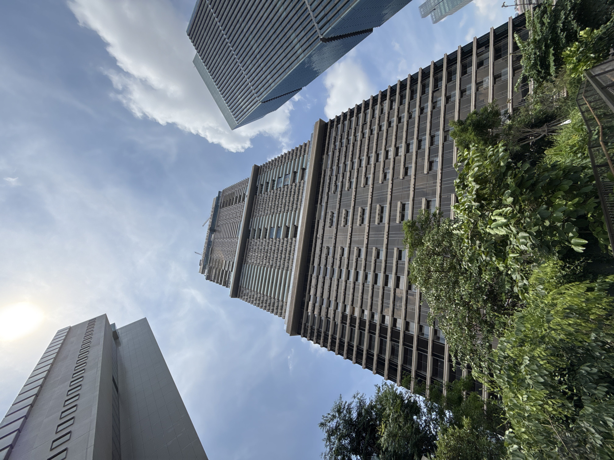 The image shows a low-angle view of skyscrapers in Bangkok, Thailand, against a backdrop of a blue sky with scattered clouds. Two prominent high-rise buildings dominate the frame; the building on the right is taller and has a facade with dark vertical elements, partially obscured by green foliage from trees in the foreground. The building in the upper left has a reflective, horizontally-oriented facade. A third building is visible in the lower-left corner, appearing more solid and less reflective than the others. A glimpse of sunlight peeks between the buildings, creating a bright spot in the frame.