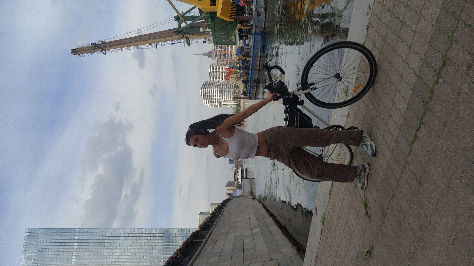 A full shot shows a woman with dark hair tied in a ponytail, wearing a white tank top and brown pants, standing next to a bicycle on a paved walkway. She is positioned on the right side of the frame, leaning her left arm on the handlebars of the bicycle. Her gaze is directed towards the right.
Behind her, a river or canal is visible, with murky water reflecting the cloudy sky. To the left of the water, a stone embankment lined with greenery runs along the walkway. In the background, a tall, modern glass skyscraper dominates the left side of the cityscape. On the right side of the frame, a large industrial crane with a yellow boom and a green base is situated on a barge in the water. Various other barges and buildings are visible in the distance, suggesting an urban industrial waterfront. The sky is overcast with scattered clouds.