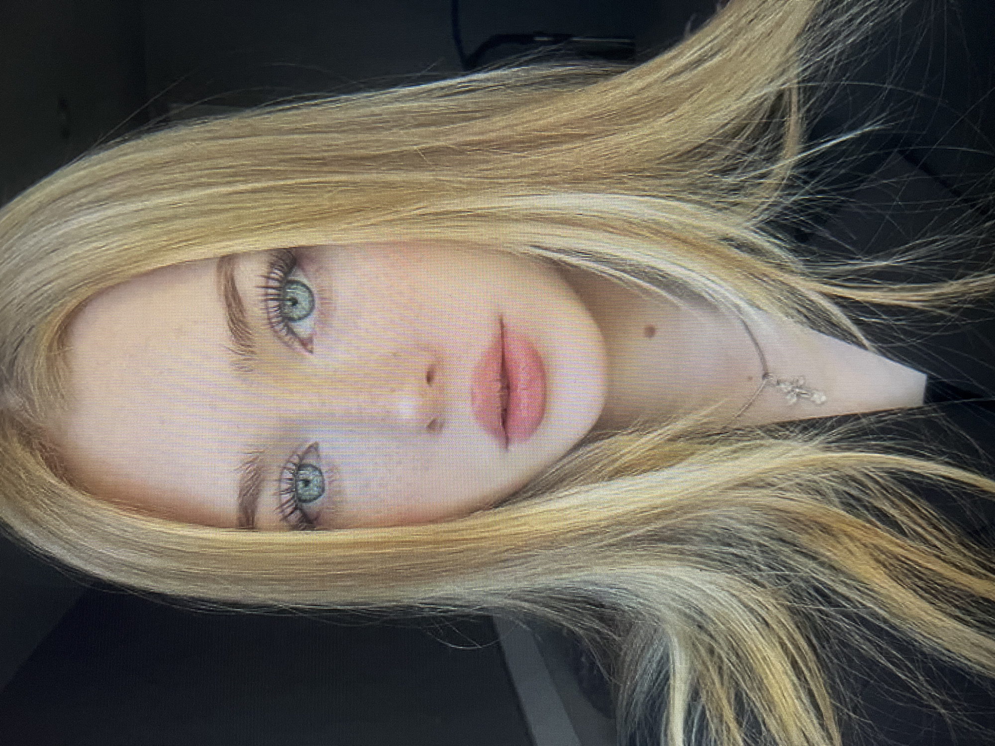 A close-up, head-and-shoulders portrait of a young Caucasian woman with long, straight, blonde hair. She has fair skin with light freckles across her nose and cheeks. Her eyes are a striking blue-green, and she has long, dark eyelashes. Her lips are full and appear to be tinted with a pale pink lipstick. She is wearing a dark jacket or blazer and a delicate silver necklace with a cross pendant. The background is dark and out of focus. The image appears to be a digital capture, possibly from a screen, given the visible pixelation. There is no discernible connection to Qarağandı city administration, Kazakhstan.