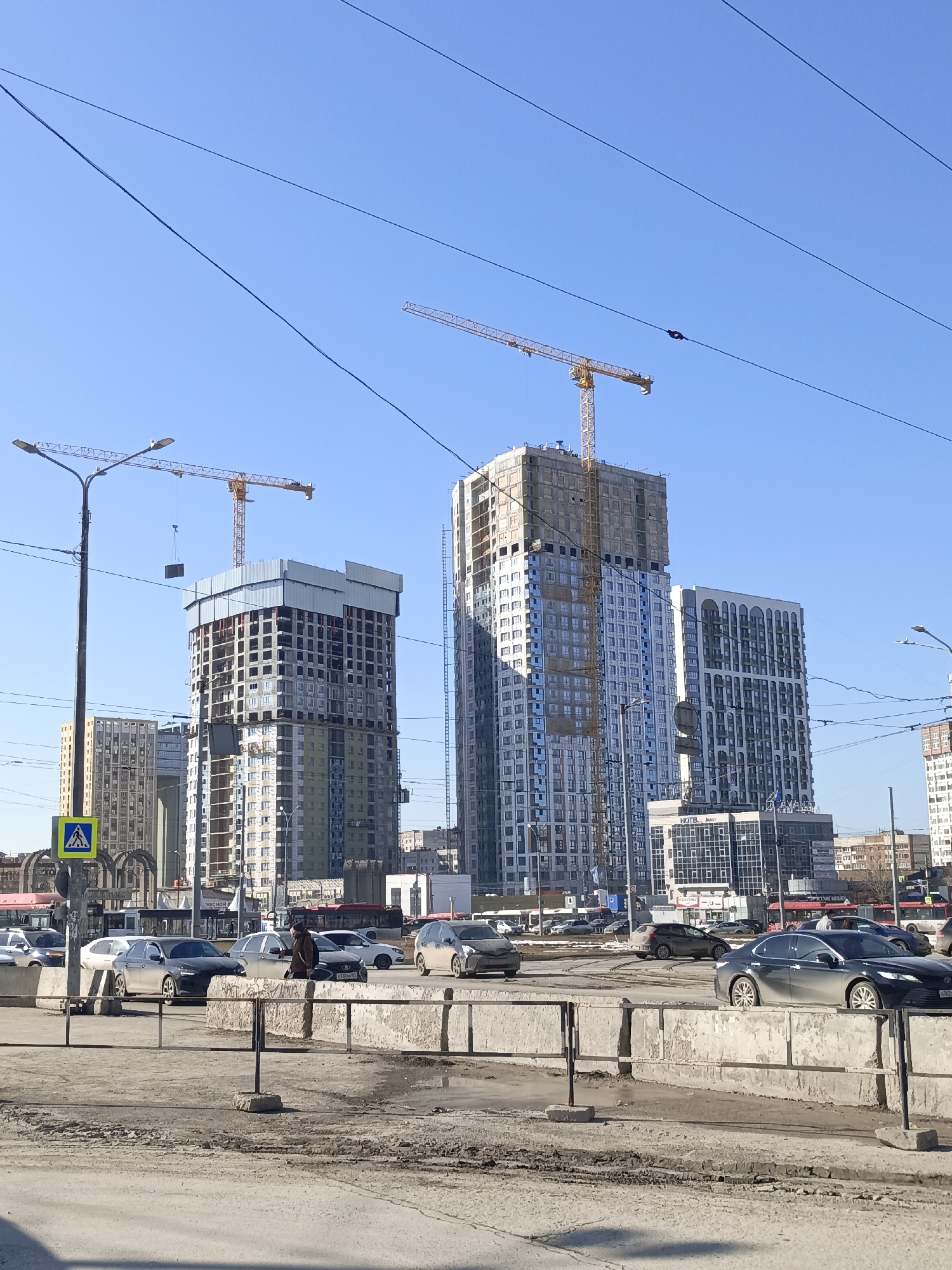 The image shows a cityscape in Perm, Russia, featuring several high-rise buildings, including some under construction with visible cranes. The foreground contains a busy street with cars, pedestrian barriers, and signs. The sky is clear blue. Several pedestrians are visible on the edge of the road. There are power lines stretching across the sky.