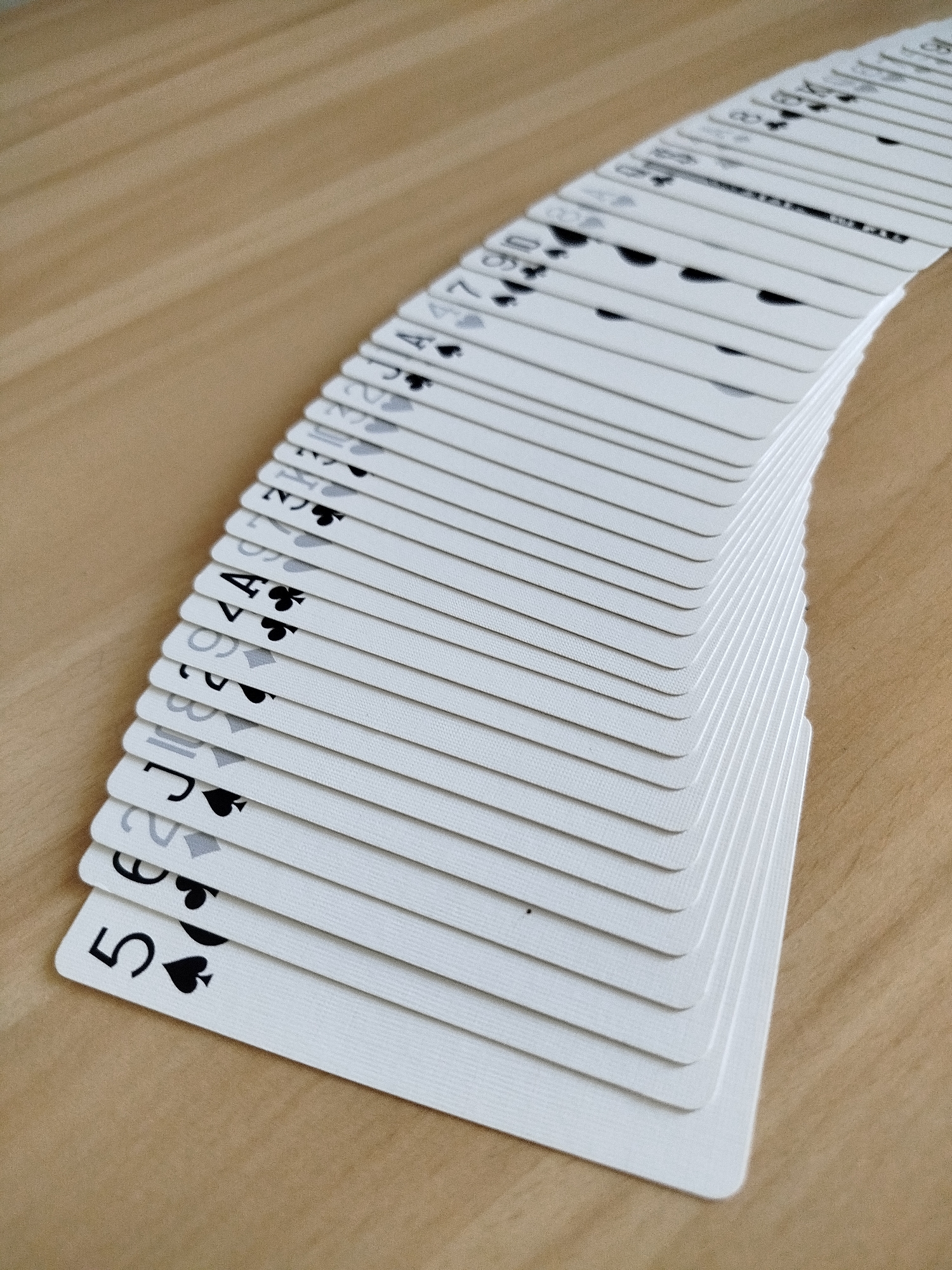 A close-up, high-angle shot showcases a fanned-out deck of white playing cards arranged diagonally on a light brown wooden surface. The cards are partially visible from the top, revealing their numerical and suit markings in black. The deck starts with the '5 of Spades' at the bottom left and fans out towards the top right, with cards overlapping and progressively obscuring the ones beneath. The wooden surface exhibits a fine grain pattern. The scene is indoors, as suggested by the lighting and the nature of the object. There is no indication of the city of Varna, Bulgaria, or any urban environment.