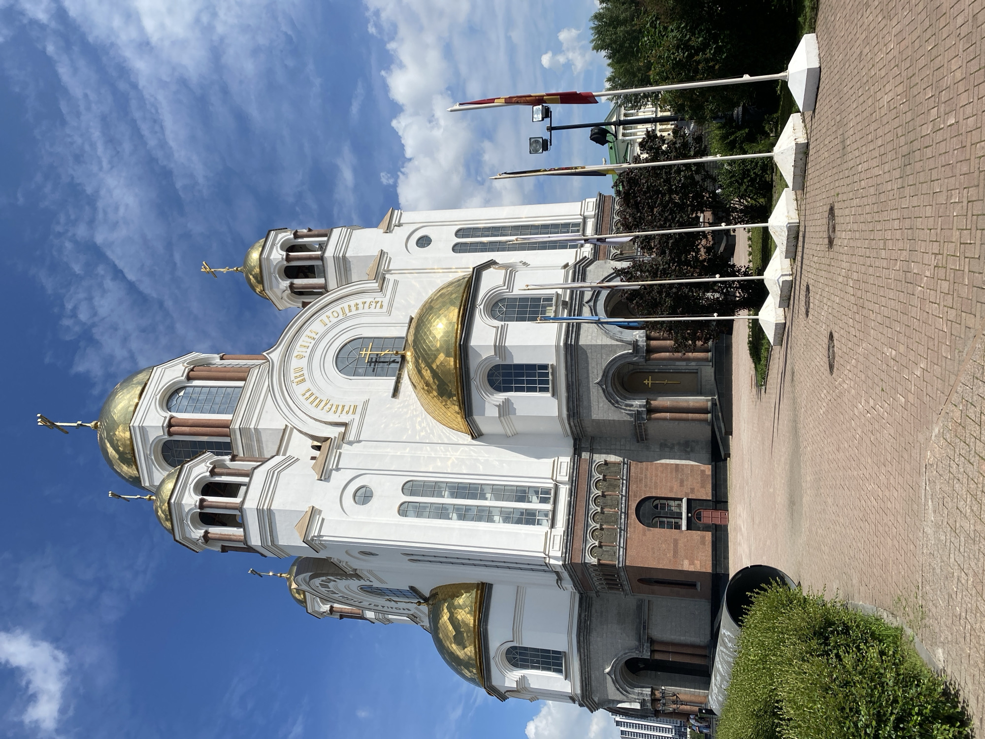 The image shows the Church of All Saints in Yekaterinburg, Russia, on a sunny day with a partly cloudy sky. The church is white with golden domes and features prominent crosses. In front of the church, several flags are displayed on white poles, standing on square bases. To the left of the church, a large dark pipe lies on the ground next to a neatly trimmed green hedge. The ground in the foreground is paved with red bricks in a herringbone pattern. In the background, trees and other buildings are visible, including what appears to be a modern high-rise to the far left. There are no visible people in the immediate foreground of the image.