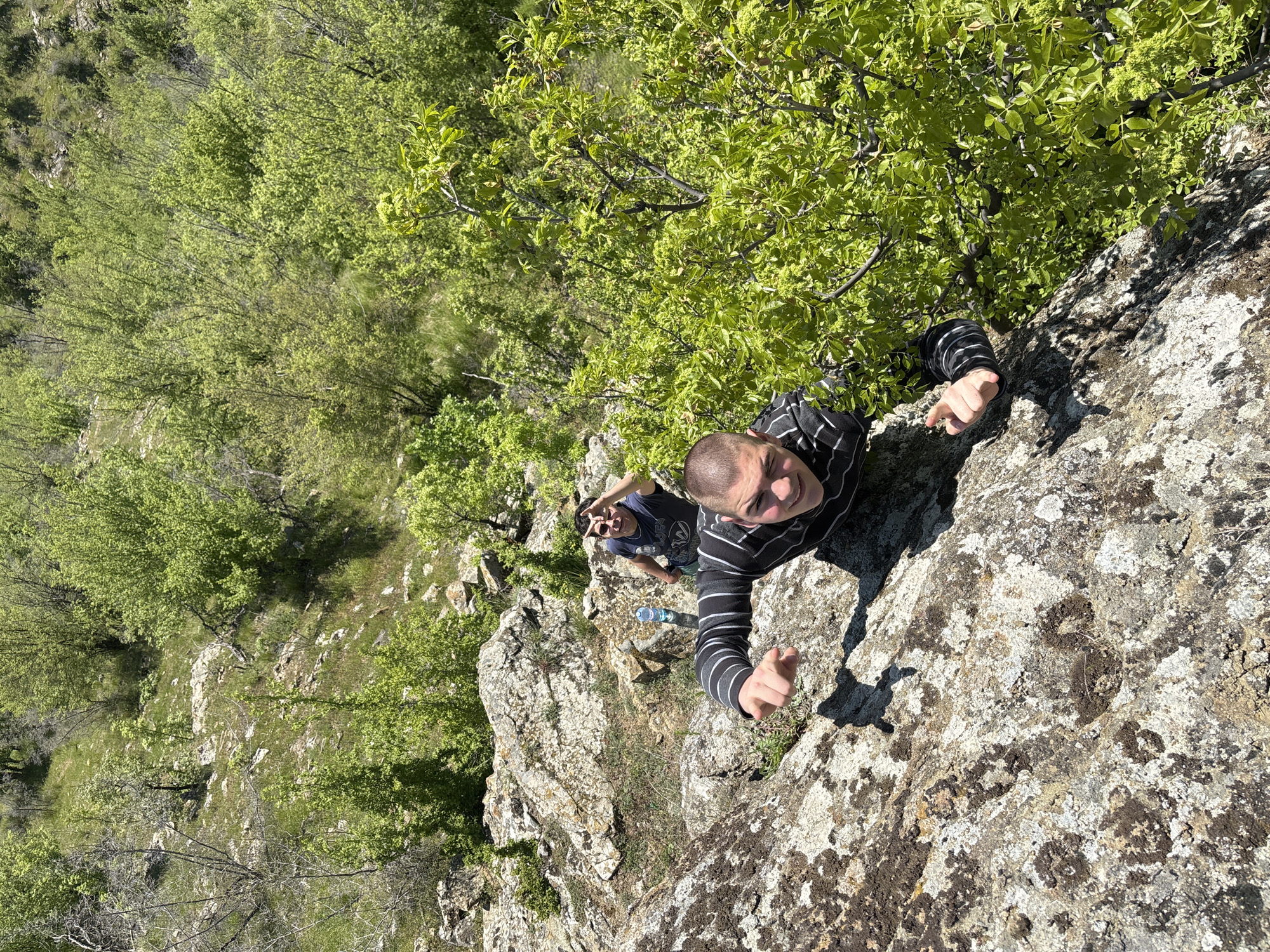 The photograph depicts two men on a rocky outcrop in what appears to be a natural setting, possibly near the city of Goritsa, Bulgaria. The man in the foreground is wearing a black and white striped long-sleeved shirt and is extending his arms towards the camera. The other man is wearing a dark t-shirt and has his fingers up in a 'peace' sign. A clear water bottle is on the rocks nearby. The surrounding environment includes green trees and other rocks with some vegetation.