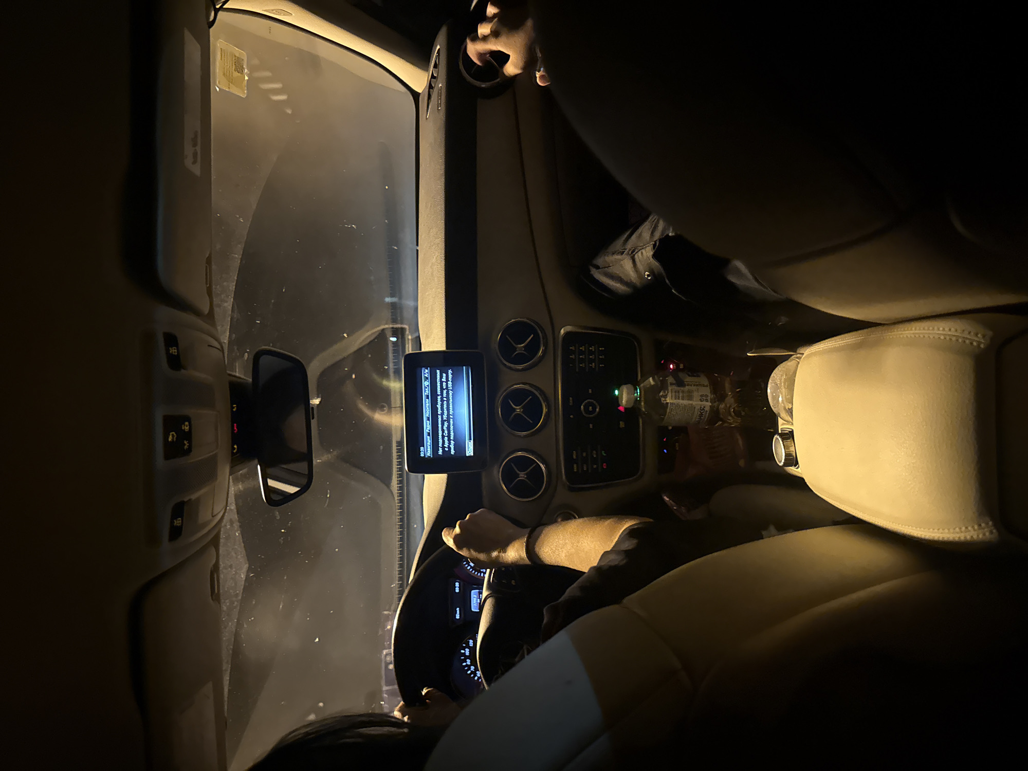 The image shows the interior of a car at night. The driver is visible in the foreground, holding the steering wheel with both hands. A passenger is visible in the front passenger seat. The car's dashboard is visible, with a navigation screen displaying a message in Russian. Other details include the rearview mirror, air vents, and the car's seats. The location cannot be determined from the image.