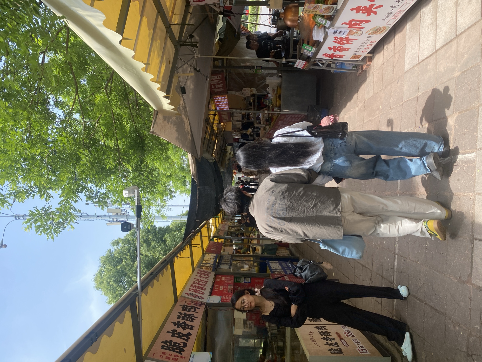 Here is a detailed description of the image:
**General Impression:** The image shows a bustling street scene in what appears to be a market or commercial area. The perspective is from a slightly elevated position on the ground.
**Location:** Based on the signage, the presence of Chinese script, and general architectural style, it is highly probable that this is a street in the Shapingba District of China.
**People:**
* Several people are visible, including three individuals in the foreground walking or standing on the street.
* One person is wearing a black outfit with glasses and appears to be posing for the camera or looking directly at the viewer.
* Two people are walking ahead, one in blue jeans and a white shirt and the other in off-white pants and a brown jacket.
* There are other people in the background, likely customers and vendors.
**Objects:**
* The street is lined with shops and stalls, as evidenced by the awnings and signage.
* There are visible signs written in Chinese characters advertising food or goods, including something related to "Crispy Duck."
* There's also a food preparation stall with a cooking station, and some individuals appear to be working at these stations.
* Miscellaneous items related to street vending, such as food containers and cooking equipment, are visible.
* The street surface is paved.
**Actions and Interactions:**
* People are walking along the street, likely shopping or commuting.
* Vendors seem to be engaged in selling goods or preparing food.
* There is a potential interaction with the photographer, as one person is looking directly at the camera.
**Environment:**
* The scene takes place outdoors on a sunny day.
* The atmosphere appears to be lively and crowded.
* Trees are visible, suggesting that the street may be lined with greenery.
* The general impression is that of a thriving commercial district.