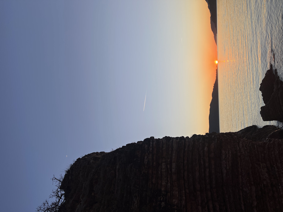 The image displays a coastal scene at sunset in Rafailovići, Montenegro. A large, dark, multi-layered rock cliff occupies the left side of the frame, exhibiting a rough, stratified texture. Sparse, dark, leafless vegetation is visible along the top edge of the cliff. In the upper left sky, a faint crescent moon is discernible. The upper sky is a clear, dark blue, transitioning downwards into a lighter blue, then an orange and yellow gradient towards the horizon. A distinct white aircraft contrail extends horizontally across the middle of the sky, with another fainter contrail positioned higher.
The lower right portion of the image features a calm body of water, likely the Adriatic Sea, reflecting the orange and yellow light of the setting sun. The sun itself, a bright orange disc, is visible low on the horizon, partially obscured by distant, silhouetted landmasses or hills that frame the central view of the sunset. At the base of the main cliff, several smaller, dark rock formations are partially submerged in the water. No individuals, animals, or specific man-made structures beyond the aircraft contrails are present in the scene.