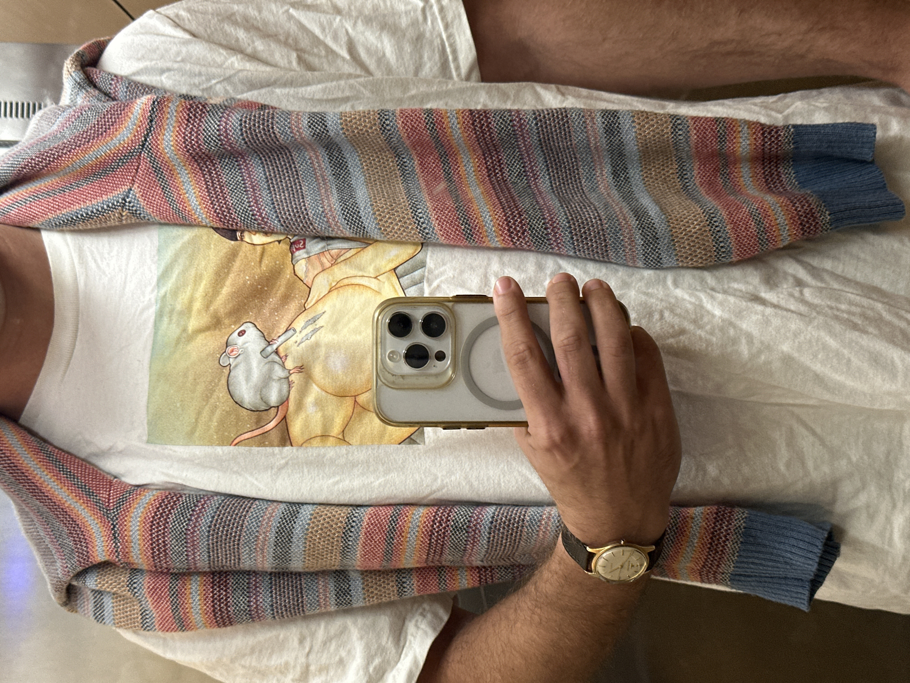 An overhead shot shows a person in a white t-shirt. The shirt has a graphic image of a woman and a mouse. A colorful striped sweater with blue ribbed cuffs is draped over the shirt. A hand holds a smartphone with a clear case over the center of the shirt. A gold watch with a dark leather band is worn on the wrist of the hand holding the phone.