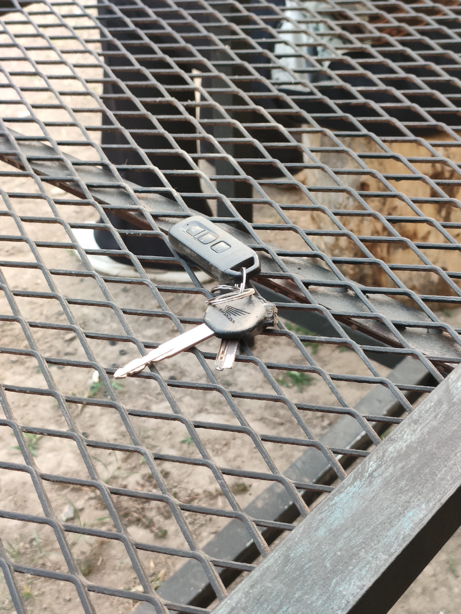 A set of keys and a key fob rest on a metal grate outdoors. The key fob is black with three buttons and appears to be for a Honda vehicle, as indicated by the embossed logo on the key. The keys are silver and have a metallic sheen. The grate is made of diamond-shaped mesh and is dark grey. The background consists of dirt and dry grass, suggesting an outdoor, possibly urban or rural, environment. The lighting is natural, indicating daylight. The scene does not depict any specific location within Banguntapan, Indonesia, other than the general outdoor setting. No people or actions are visible.