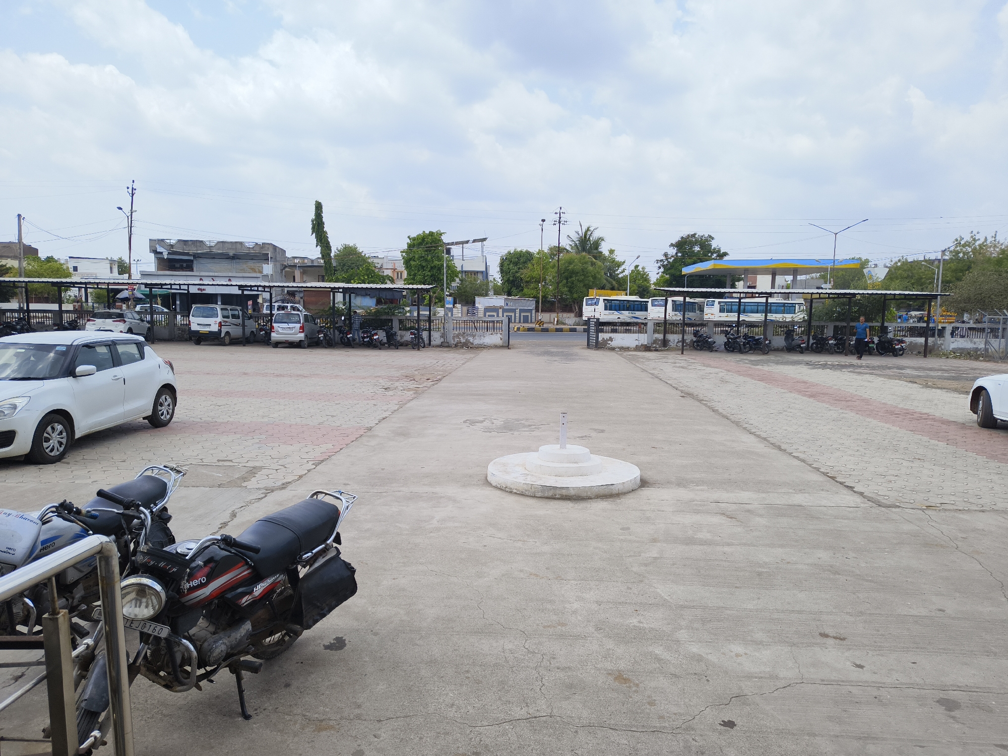 The image depicts a parking area in Dhandhuka, India, under an overcast sky. The foreground is dominated by a concrete expanse with a circular, tiered, white structure in the center. A motorcycle, a black "Hero" model, is parked near the left edge of the frame, partially obscuring another motorcycle. A white Suzuki car is visible on the left. Rows of parked motorcycles are seen under sheltered structures further back. In the background, there are buildings and a blue and yellow gas station. A bus is parked near the gas station. Several cars and vans are visible in the distance near the buildings. The ground surface is a mix of concrete and brick-like paving.