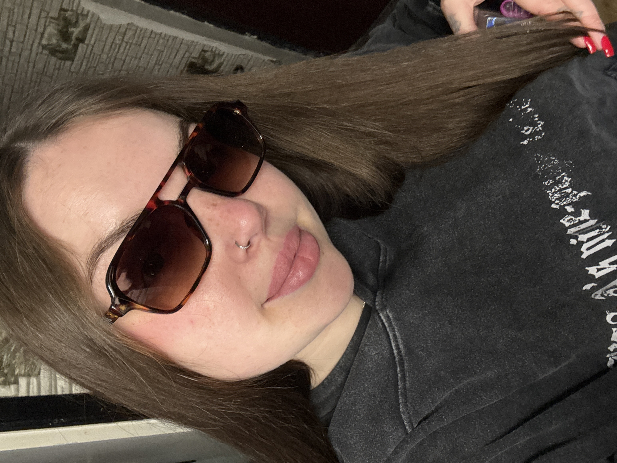 A woman lies at an angle in a dark setting, wearing brown sunglasses with a hexagonal frame. She has long, straight, brown hair and a nose ring. Her lips are full and appear to be light pink. She is wearing a dark gray sweatshirt with white graphic lettering. Her hand, with red nail polish, is seen holding her hair. A corner of what appears to be a white desk or shelf is in the lower left corner. The location is unspecified.