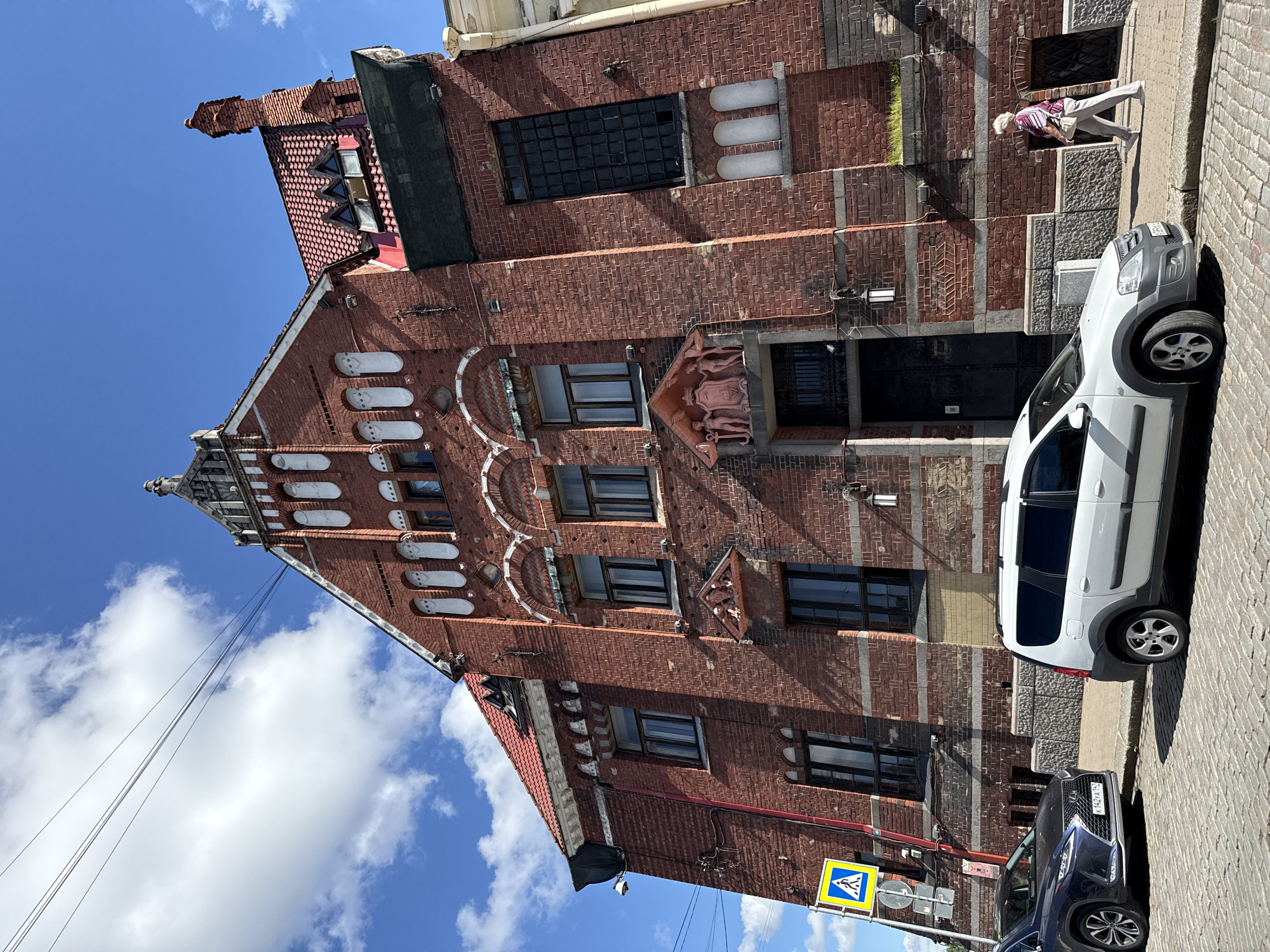 This is a low-angle, full-shot, outdoor, eye-level photograph of a red brick building in Vyborg, Russia. The building has a steeply pitched red tile roof and ornate architectural details, including arched windows with white trim, decorative sculptures above the entrance, and a pointed spire at the peak of the facade. The building's facade is a warm reddish-brown brick with horizontal bands of lighter stone. There are several windows visible on the building, some with dark panes and others with lighter reflections.
In the foreground, on the cobblestone street, are two cars: a dark blue Lexus SUV on the left and a white crossover SUV on the right. The Lexus has a license plate that reads "K 142XA 197". A pedestrian, an elderly woman wearing a striped shirt and light-colored pants, walks along the sidewalk on the right side of the frame, away from the building.
To the left of the building, a yellow and blue pedestrian crossing sign is visible on a metal pole. The sky above is a clear blue with scattered white clouds, and several thin electrical wires are strung across the upper portion of the image. The building appears to be historic, likely a commercial or residential property, given its grand architecture and placement on a city street. The overall scene suggests a bright, sunny day in a European city.