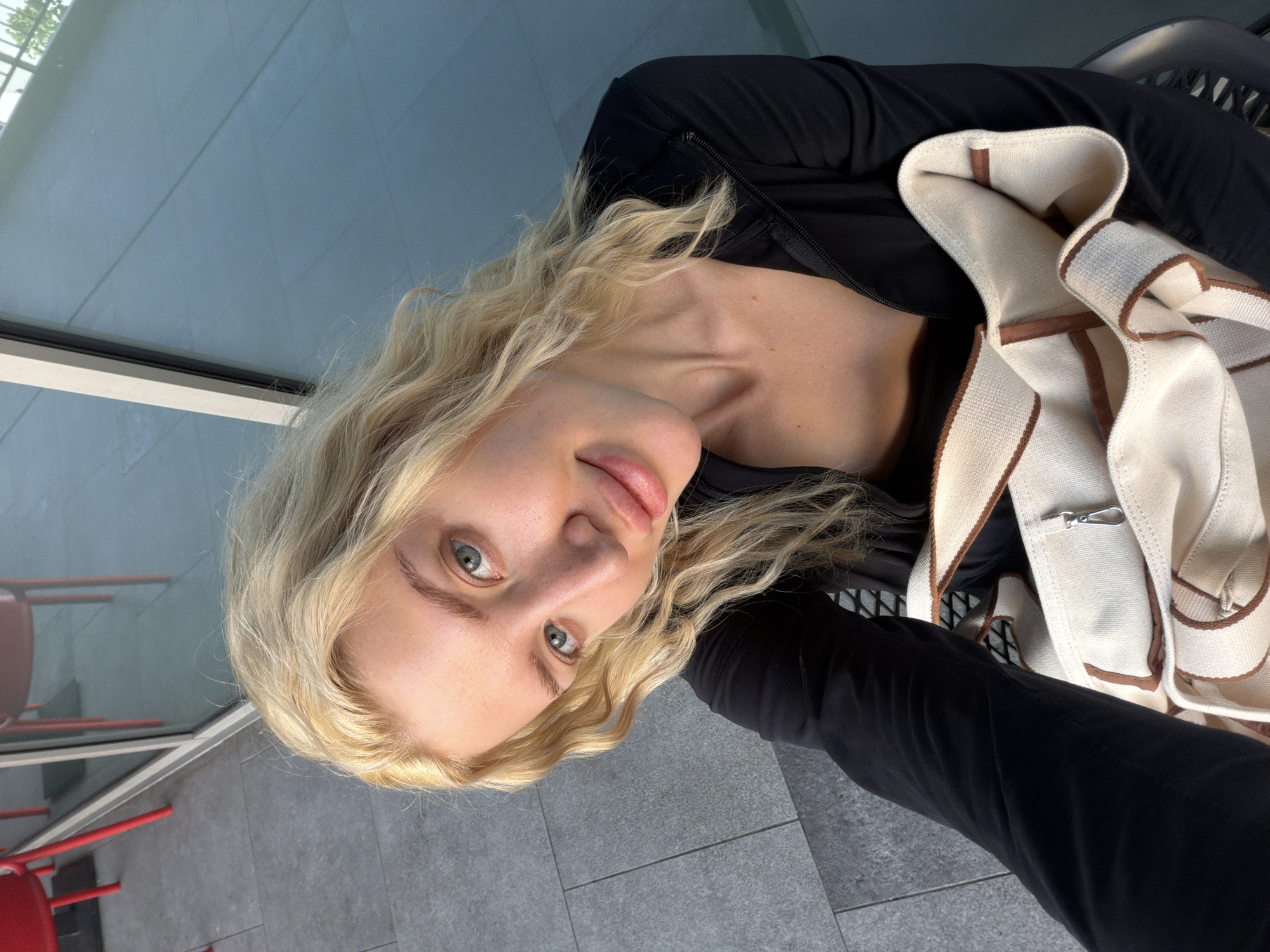 A woman with blonde wavy hair and fair skin is posing for a selfie. She is wearing a black long-sleeved top and has a beige and brown shoulder bag with a silver clasp on her chest. The background shows a reflective glass window and a gray tiled floor. Outside the window, red chairs and tables are visible, likely indicating an outdoor seating area. The scene appears to be in an urban setting, potentially a cafe or restaurant in Belgrade, Serbia, considering the city was mentioned.