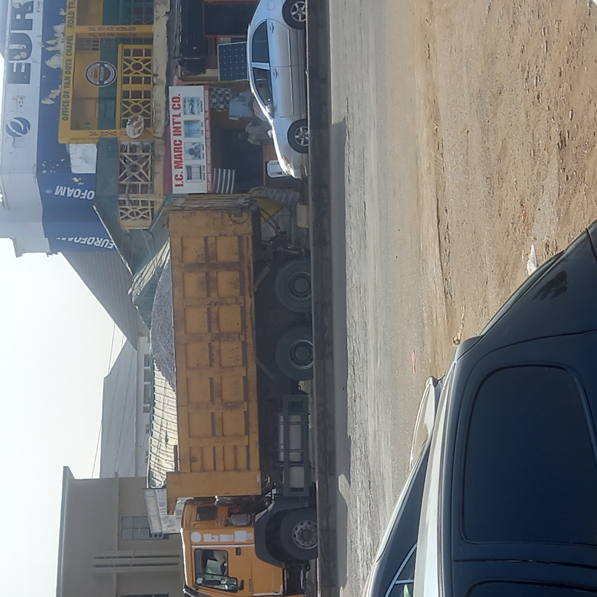 The image shows a yellow dump truck loaded with what appears to be gravel, parked on a street. Next to the truck are two cars: one silver and one dark blue. The buildings in the background have signs for "EUROFOAM" and "I.C. MARC INT'L CO." The street surface is paved. Based on the presence of "OFFICE OF YAN DOYA CHAPEL" and "ROAD TRA" on a sign, the image is likely in Dadi Riba, Nigeria.