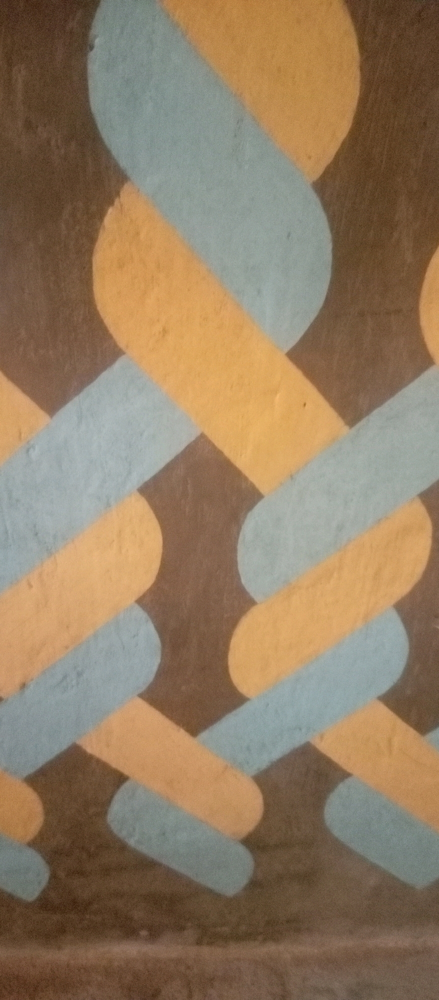 The image shows a close-up of a wall with a painted pattern. The pattern consists of interweaving bands of light blue and yellow against a dark brown background. The bands are rounded at the ends and create a woven or chain-like effect. The texture of the wall appears rough, suggesting it might be concrete or stucco. The lighting is somewhat dim, casting soft shadows and highlighting the texture of the paint and the wall. There are no people, signs, or discernible landmarks visible that would indicate a specific location within a city. The image is a detail shot of the decorative painting.