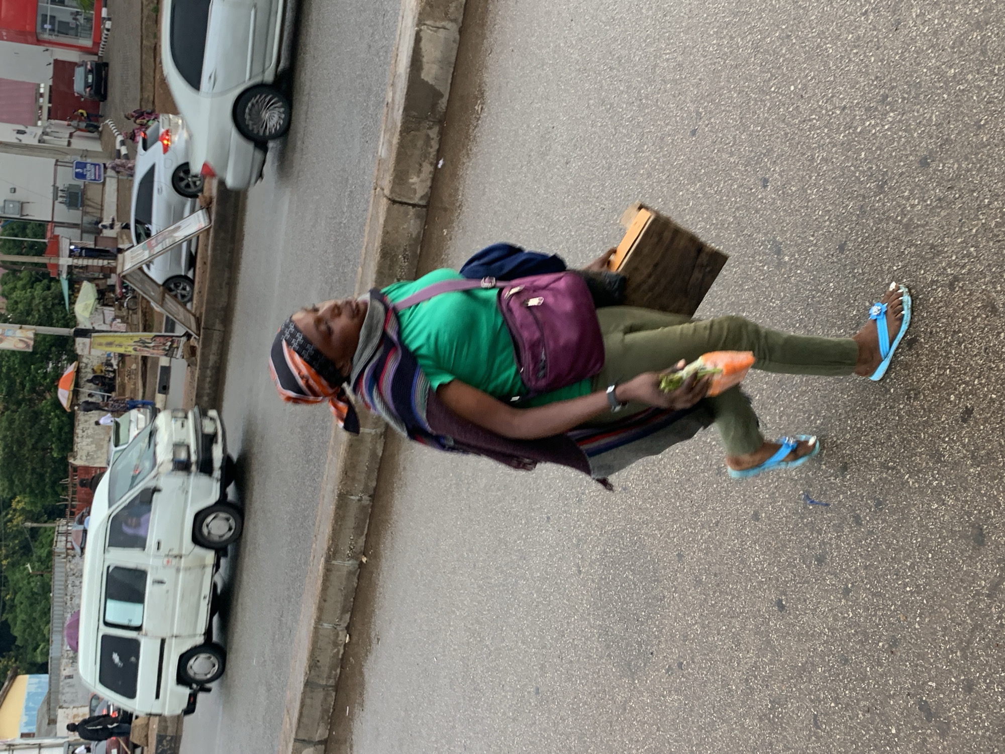A medium shot shows a woman walking on a wet asphalt road in Jos, Nigeria, possibly during or after rainfall. She is wearing a green t-shirt, olive green pants, and blue flip-flops adorned with a flower. A colorful striped scarf is wrapped around her head and shoulders, and a purple shoulder bag is slung across her body. She carries a wooden box in her right hand and a bunch of carrots in her left. Her gaze is directed forward, suggesting a determined stride. In the background, a white minivan, several cars, and a few pedestrians are visible, along with buildings and street signs. The overall impression is of a bustling urban environment, with the woman being the central focus of the image as she navigates the street.