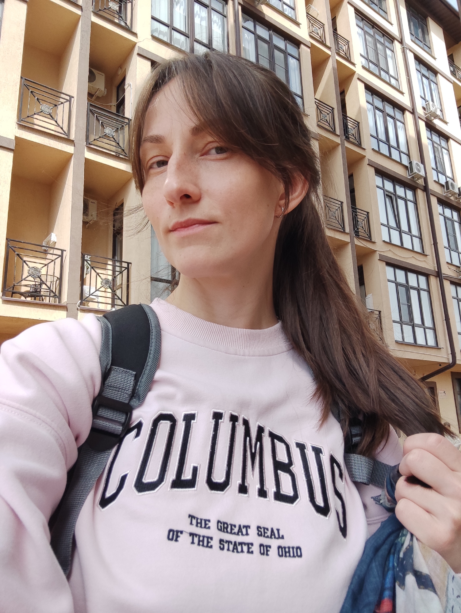 A young woman stands in front of a multi-story apartment building, likely in Sochi, Russia. The building has a light tan facade with dark-framed windows and balconies with wrought iron railings. The woman wears a pink sweatshirt emblazoned with "COLUMBUS / THE GREAT SEAL / OF THE STATE OF OHIO". She has dark hair pulled back in a ponytail and a black backpack. She is holding a piece of fabric in her right hand. Air conditioning units are visible on the exterior walls of the building.