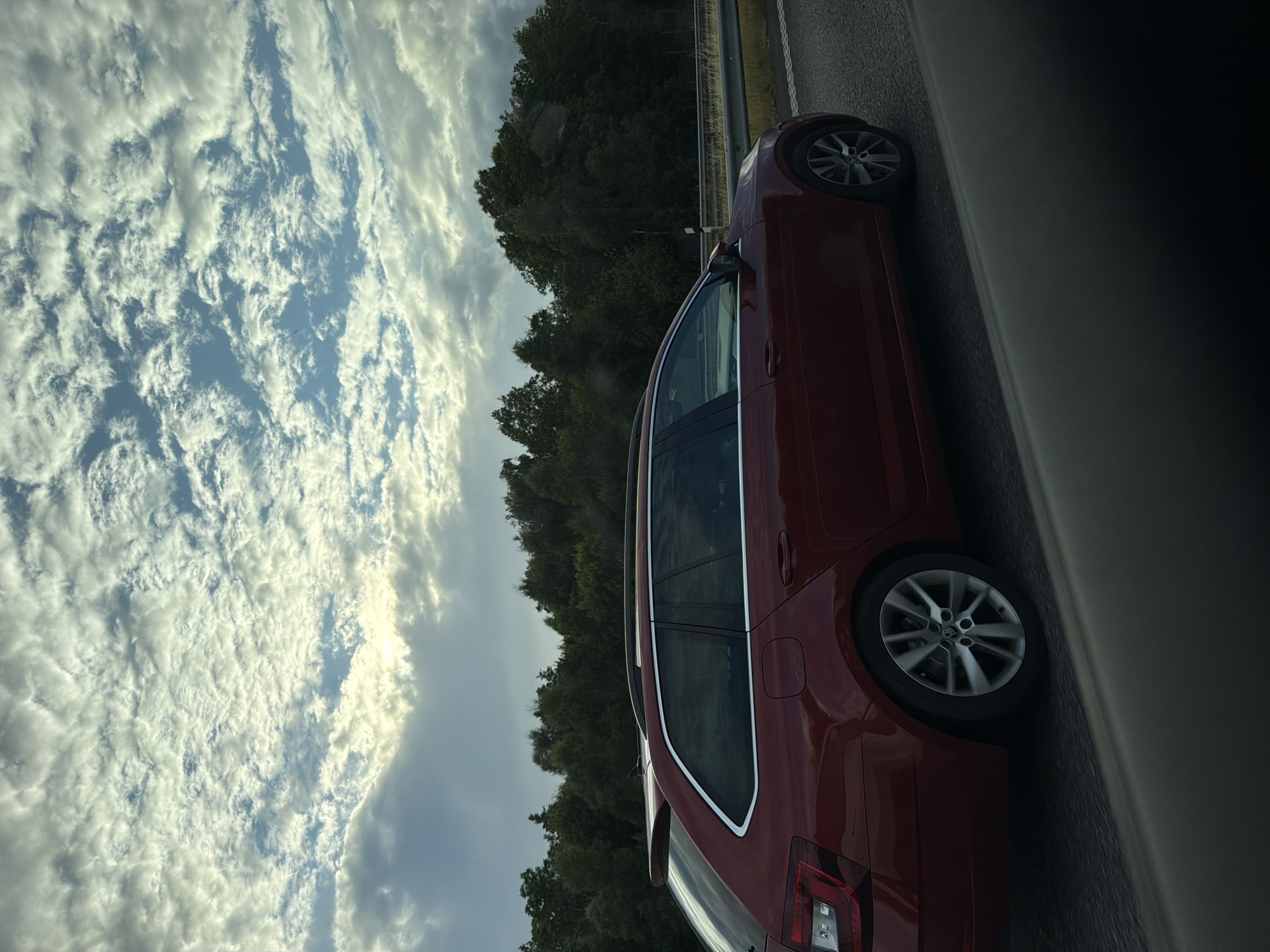 A dark red station wagon is captured in a side profile, traveling on a paved road. The car is a Skoda Octavia, indicated by the logo on the wheel. It has a silver roof rack and silver trim around the windows. The car's rear light cluster and alloy wheels are visible. The background consists of a dense line of dark green trees, suggesting a rural or suburban setting. Above the trees, a sky filled with scattered, puffy white clouds is visible, with patches of blue sky peeking through. The lighting suggests it is daytime, possibly late afternoon, given the soft sunlight filtering through the clouds. The image appears to have been taken from another vehicle, as indicated by the blurred foreground elements. The location is Åskloster, Sweden.