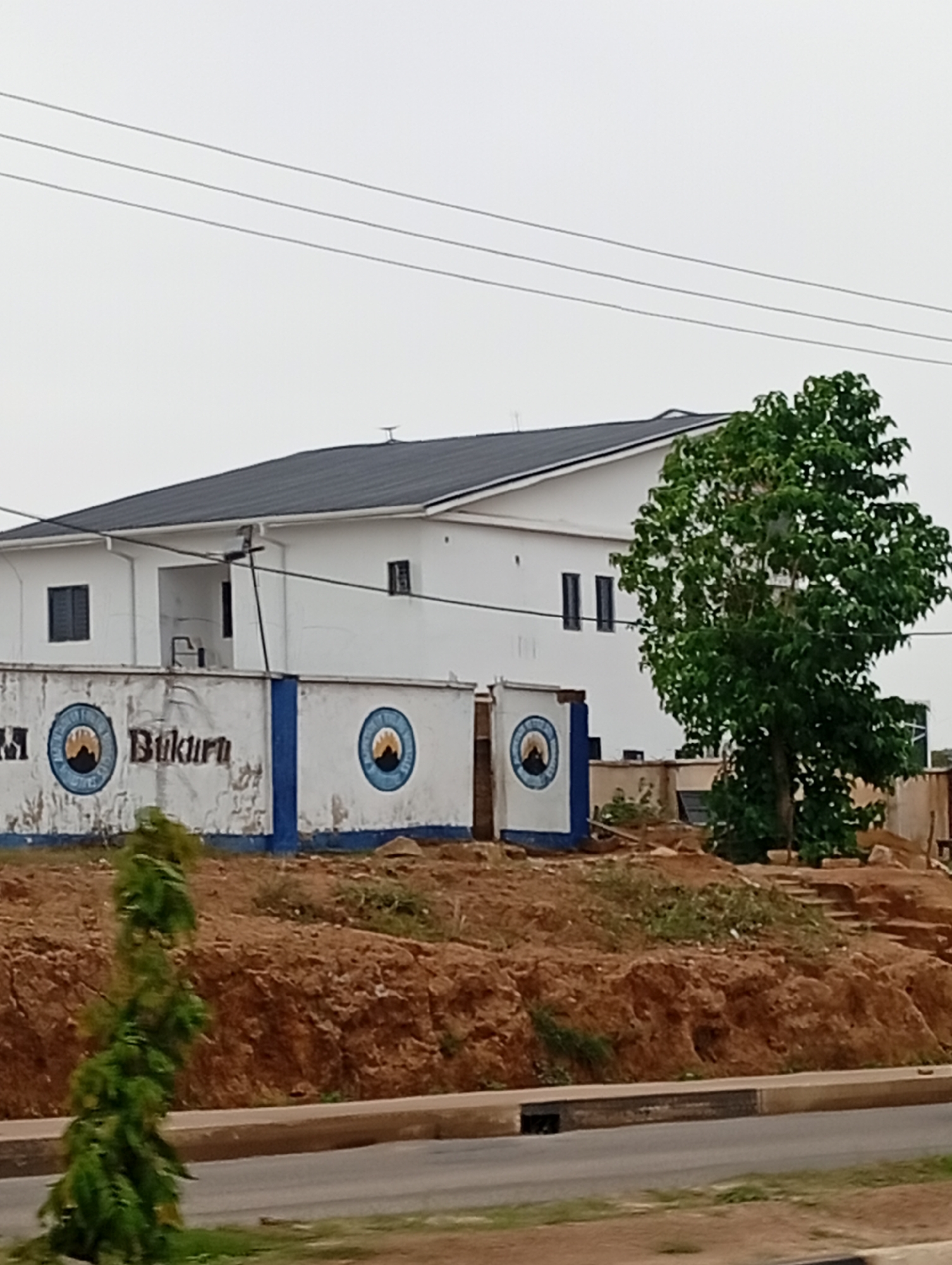 The image shows a white, two-story building with a dark-colored roof. The building is situated on a slight incline, with a retaining wall in front of it. The retaining wall has blue and white markings and appears to have emblems or logos. Text reading "Bukuru" is visible on the wall. There is a large green tree to the right of the building. In the foreground, a road is visible, and to the left of the building, a small green plant is growing. The sky is overcast. The scene appears to be in an urban or suburban area, given the presence of the road and building. The provided text "Gura Top, Nigeria" indicates the location.