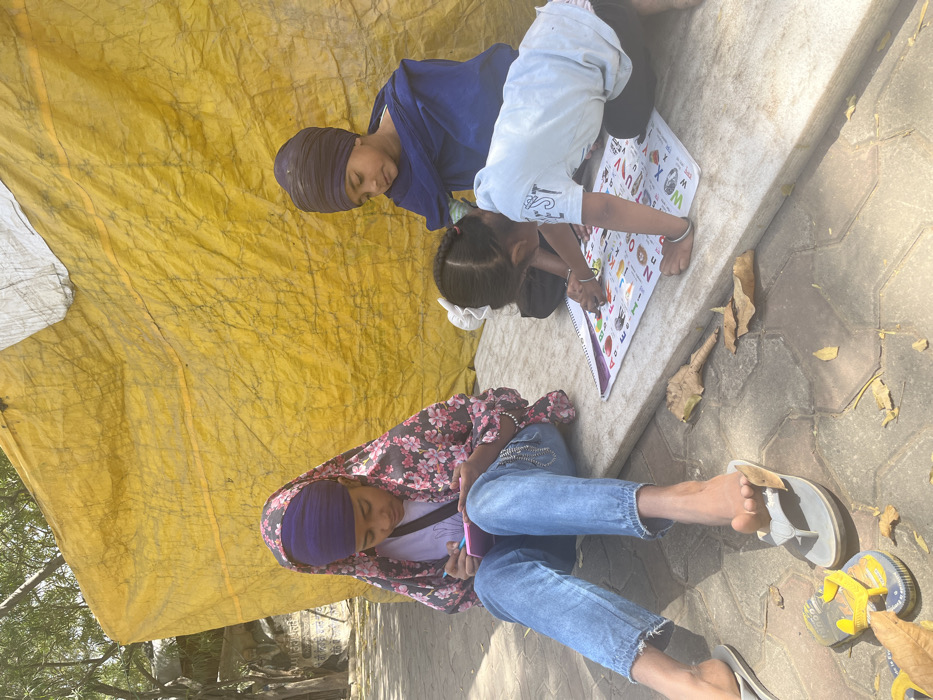 The scene takes place outside in Sahibzada Ajit Singh Nagar, India, at the coordinates provided. Three children are sitting on a paved area in front of a yellow tarpaulin. The child on the left, wearing a purple turban and a pink floral scarf, is sitting on the ground, her legs crossed. She holds a phone in her hands. Next to her, a young girl wearing a white shirt and black pants is writing on a large alphabet chart. She is sitting with her legs crossed. A third child, wearing a blue turban and blue shirt, sits behind her, watching. Dry leaves are scattered on the ground. Sunlight illuminates the scene.