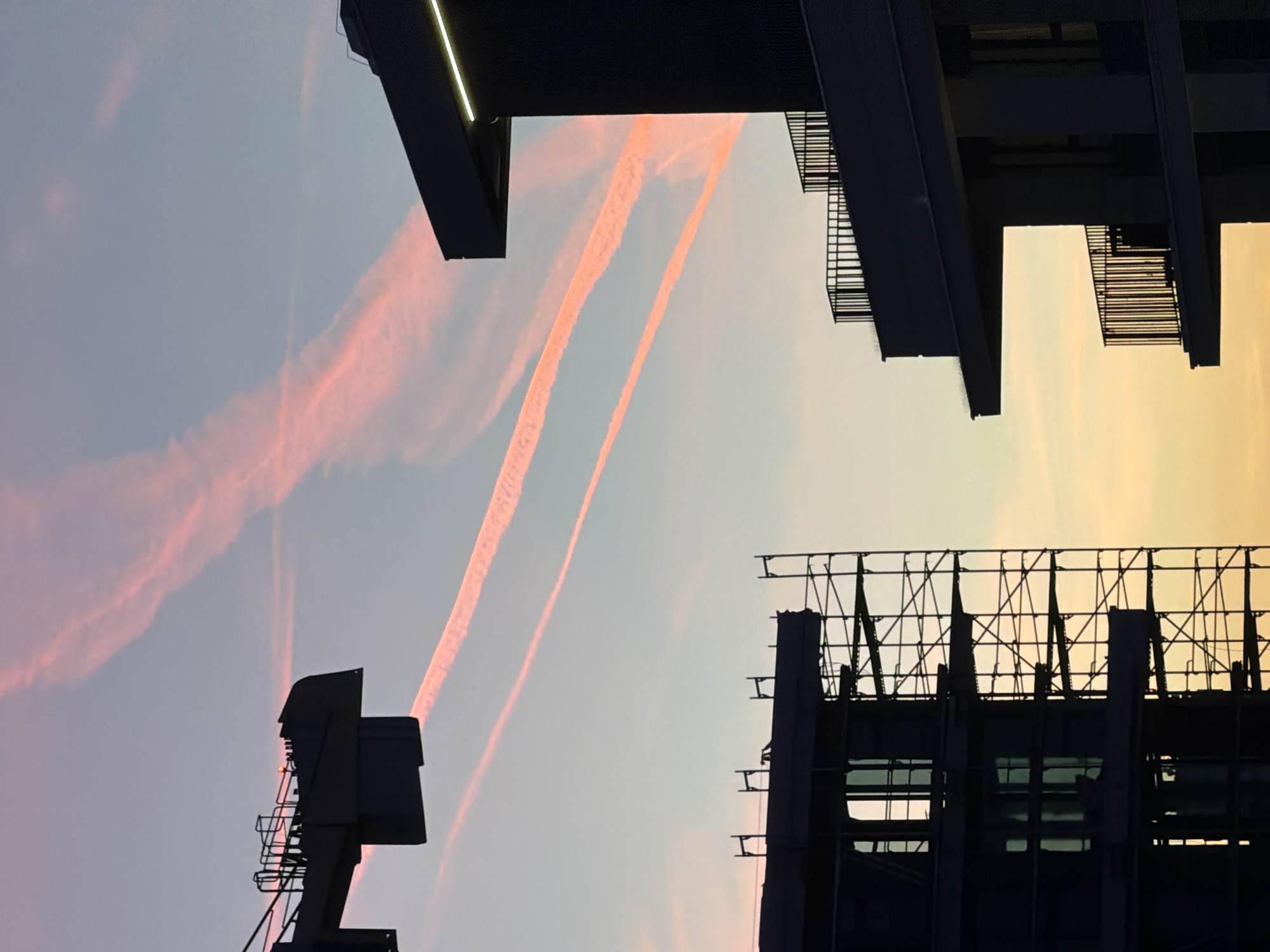 The image depicts an urban scene in Belgrade, Serbia, featuring silhouettes of buildings and infrastructure against a sky with contrails. The buildings include a modern structure with balconies and linear lighting, a billboard frame, and a crane. The sky transitions from a light blue on the left to a yellow-orange hue on the right, indicating sunset or sunrise. Several contrails are visible in the sky, appearing as pinkish streaks against the blue backdrop. The image is taken from a low angle, emphasizing the height and scale of the buildings. There are no people visible in the image.