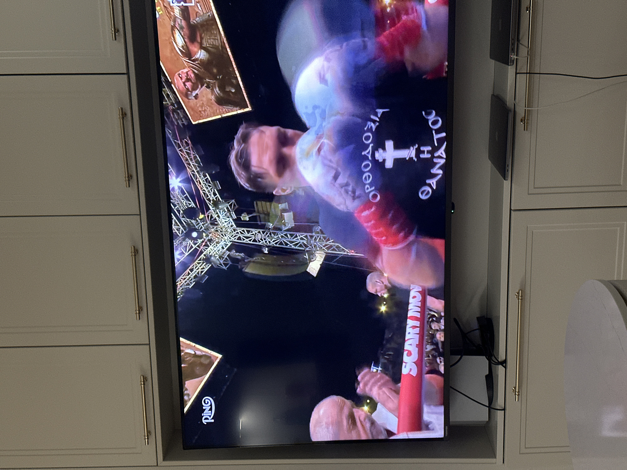 A television mounted within built-in cabinetry displays a boxing match taking place in an arena. One of the boxers is wearing a shirt with Greek text on it. The setting is Unknown, Unknown.