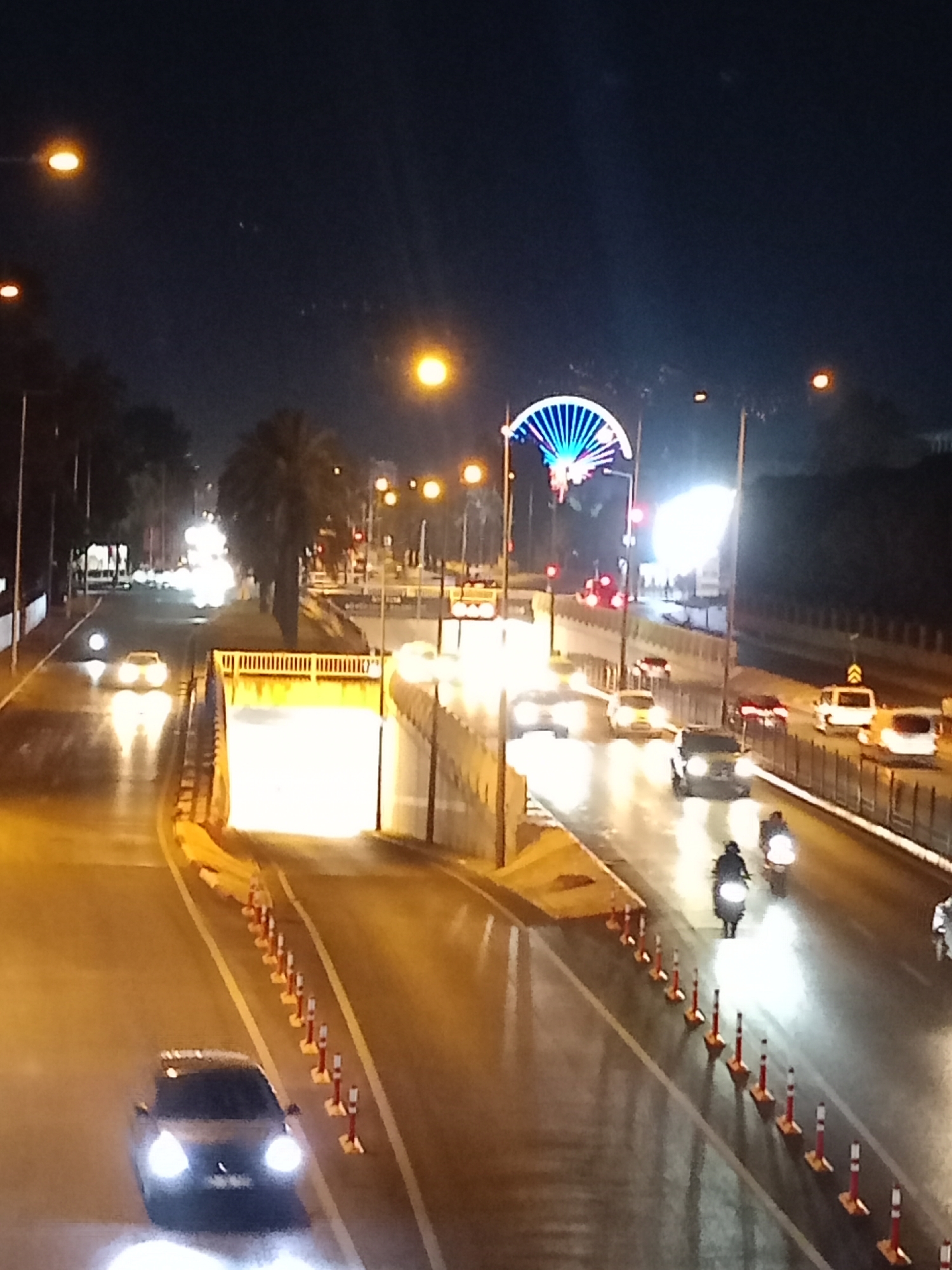 The image shows a nighttime scene of a road with several vehicles, including cars and motorcycles. The road appears to be wet, reflecting the streetlights and vehicle headlights. There is an illuminated tunnel entrance on the left side of the image, with traffic cones lining the road leading into it. In the background, there is a large, illuminated structure that resembles a Ferris wheel or a decorative archway with blue and white lights. The overall environment suggests an urban setting, possibly a highway or a major road in Antalya, Turkey, given the presence of palm trees and the architectural style of the illuminated structure. The lighting conditions are typical of a city at night, with artificial lights dominating the scene.