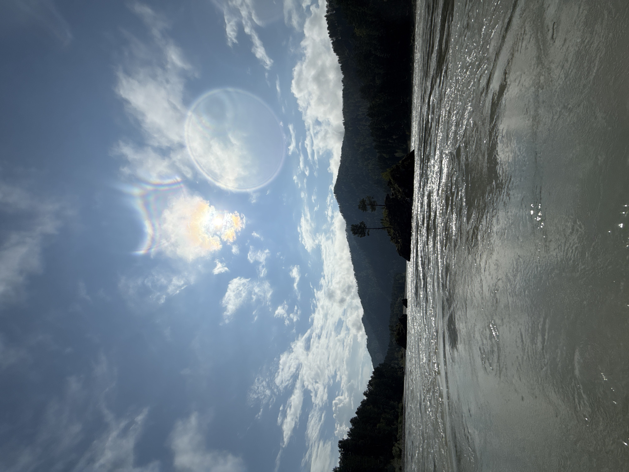 The image depicts a wide, choppy river flowing through a mountainous, forested landscape under a partly cloudy sky. The river water is a muted gray-blue, with sunlight glinting off its surface, creating a shimmering effect. In the mid-ground, a small, rocky island topped with two sparse trees stands in the river. The background is dominated by dark, tree-covered mountains that slope down towards the water. The sky is a bright blue with scattered white clouds, and there are noticeable lens flares, including a circular halo and a rainbow-colored arc, suggesting the presence of the sun. The scene is natural and serene, with no people or man-made structures immediately visible. The location is identified as Barangol, Russia.