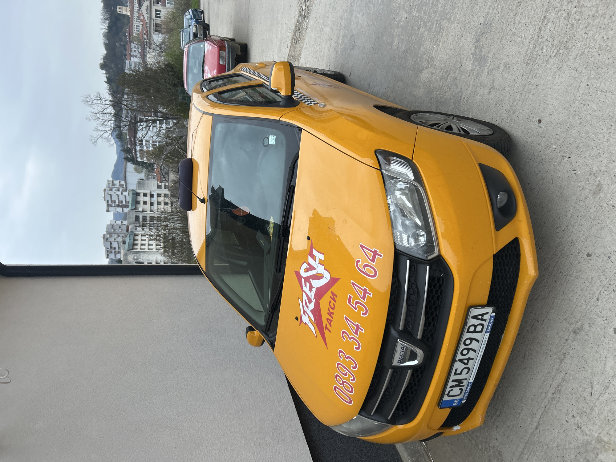 The image features a yellow Dacia taxi, parked on a concrete surface. The taxi displays the license plate "CM5499 BA" and the taxi company's branding, "FRESH ТАКСИ", along with the phone number "0893 34 54 64." The background shows a cityscape with buildings, trees, and overcast skies. Based on the taxi company's branding and the license plate, the location is likely Zlatograd, Bulgaria. Other vehicles are parked further down the road. The picture appears to be taken during the day with natural lighting.