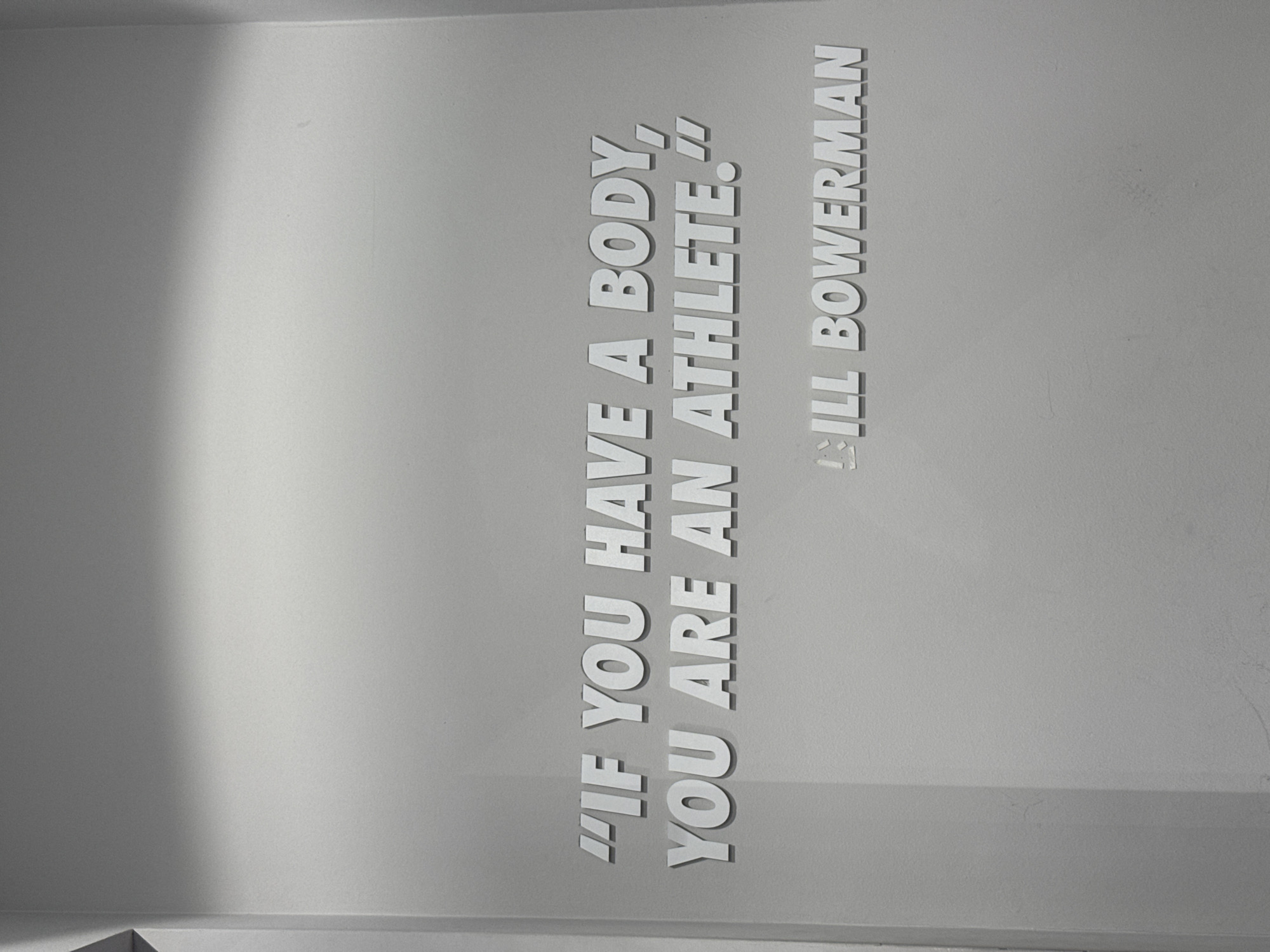 The image captures a light gray wall in an indoor setting, possibly in Sofia, Bulgaria. The wall features a motivational quote in raised, white, sans-serif capital letters, creating subtle shadows that give them a three-dimensional appearance. The text is oriented vertically, running upwards from bottom to top on the right side of the frame. A soft, diagonal shadow falls across the left portion of the wall, indicating an internal light source from the upper right. No people or activities are visible.
The prominent text on the wall reads:
"IF YOU HAVE A BODY,
YOU ARE AN ATHLETE."
BILL BOWERMAN