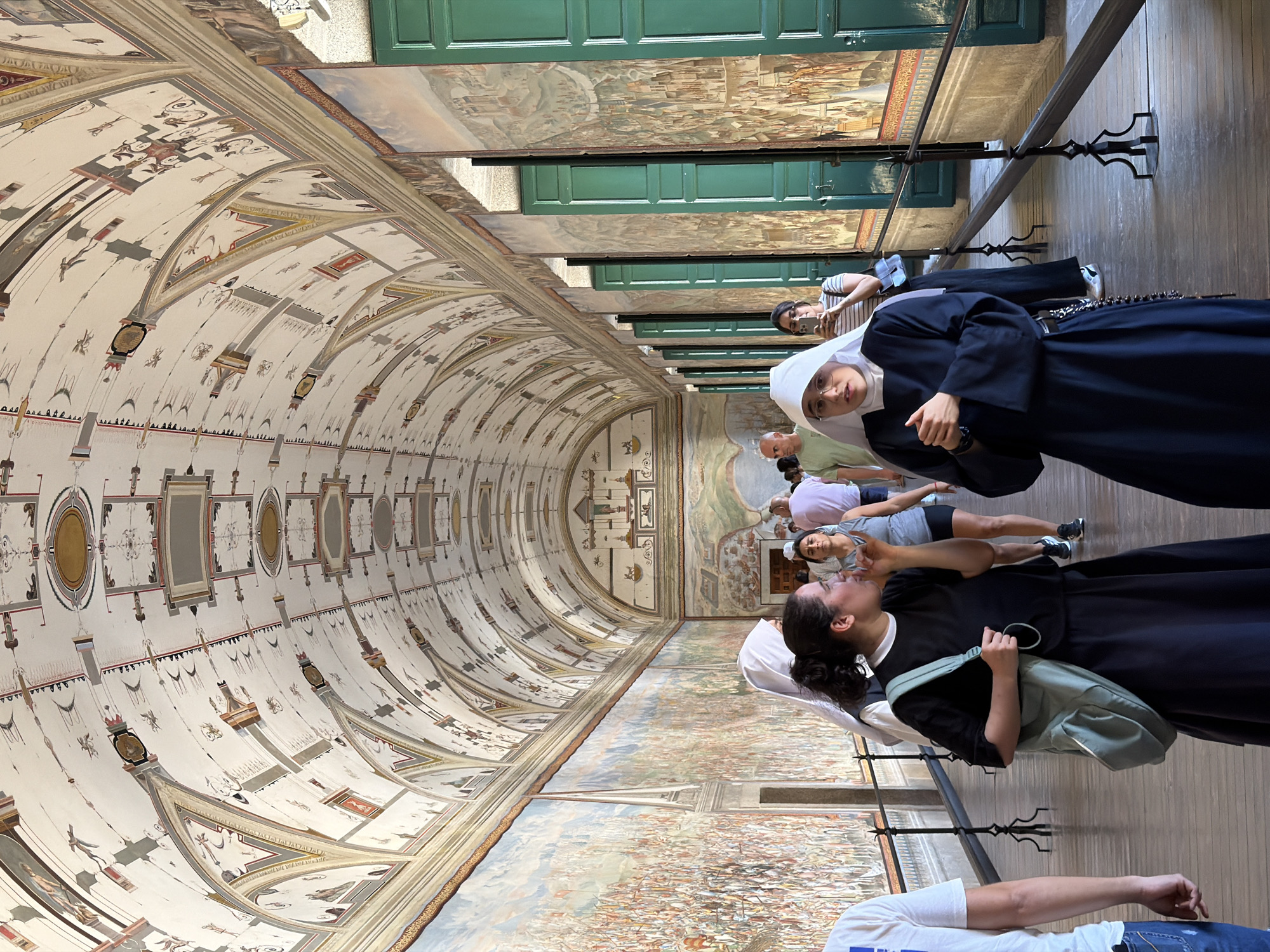 The image depicts an interior walkway within the Royal Site of San Lorenzo de El Escorial in Spain. The walkway is characterized by a vaulted ceiling adorned with intricate frescoes, featuring geometric patterns and various figures. Along the walls, large-scale paintings depict historical scenes, with a prominent mural on the left showing a multitude of people. Green shutters are visible on the right side, set within arched openings.
Several individuals are present in the walkway, appearing to be tourists or visitors. In the foreground, two women are visible. One, on the left, is wearing a black dress and a light blue shoulder bag and is gesturing with her hand. To her right and slightly behind her, a nun in a dark blue habit and white wimple is looking towards the viewer, with her hands clasped. Other people are seen further down the walkway, including a man in a light green shirt and a woman holding a smartphone. The floor is made of wooden planks, and a black metal railing runs along the right side of the walkway, separating it from the wall murals. The lighting suggests it is daytime, with natural light entering from unseen sources.