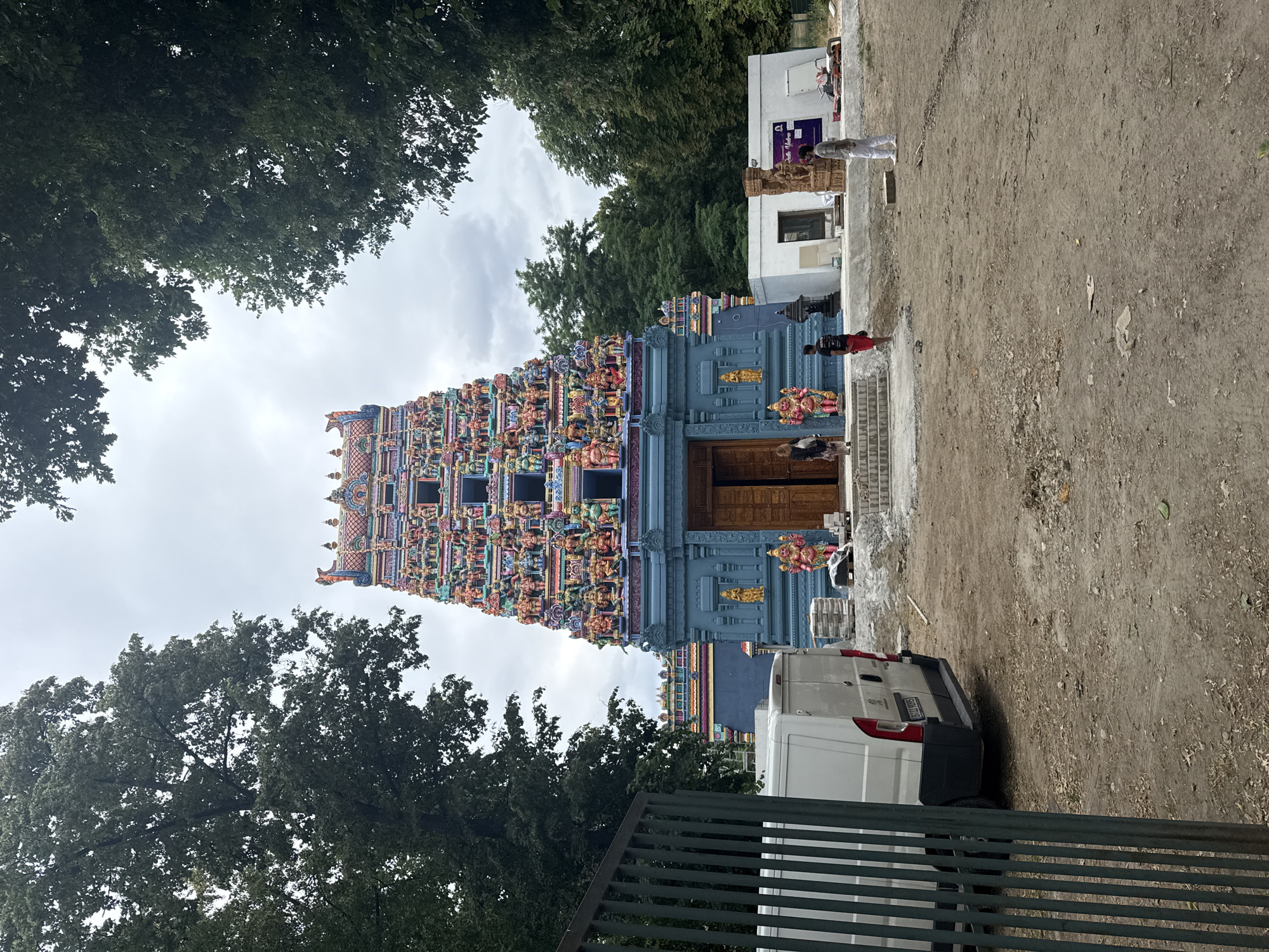 This media file features a colorful Hindu temple with intricate carvings and statues adorning its facade. The temple is a prominent subject, with its tall, multi-tiered gopuram dominating the left side of the frame. Several people are visible, including a few standing near the entrance of the temple and others in the background, some appearing to be workers. An object, a white van, is parked in the foreground on the right side.
The scene appears to be an outdoor setting in what looks like an urban or semi-urban area, with trees framing the top of the image and a dirt and gravel ground in the foreground and right side. The sky is overcast, suggesting a cloudy day. The detailed carvings on the temple indicate it is a place of religious and cultural significance. The presence of construction materials in the background might suggest ongoing work or expansion related to the temple or its surroundings.
There is a visible license plate on the white van. Additionally, some text is visible on a sign on a white building in the background, which reads "Priya Indische Kultur." The overall mood is neutral, focusing on the architectural splendor of the temple and the everyday activities around it. The time of day is unclear due to the overcast sky. The location context is Berlin, Germany, which is consistent with the presence of a diverse religious and cultural establishment.