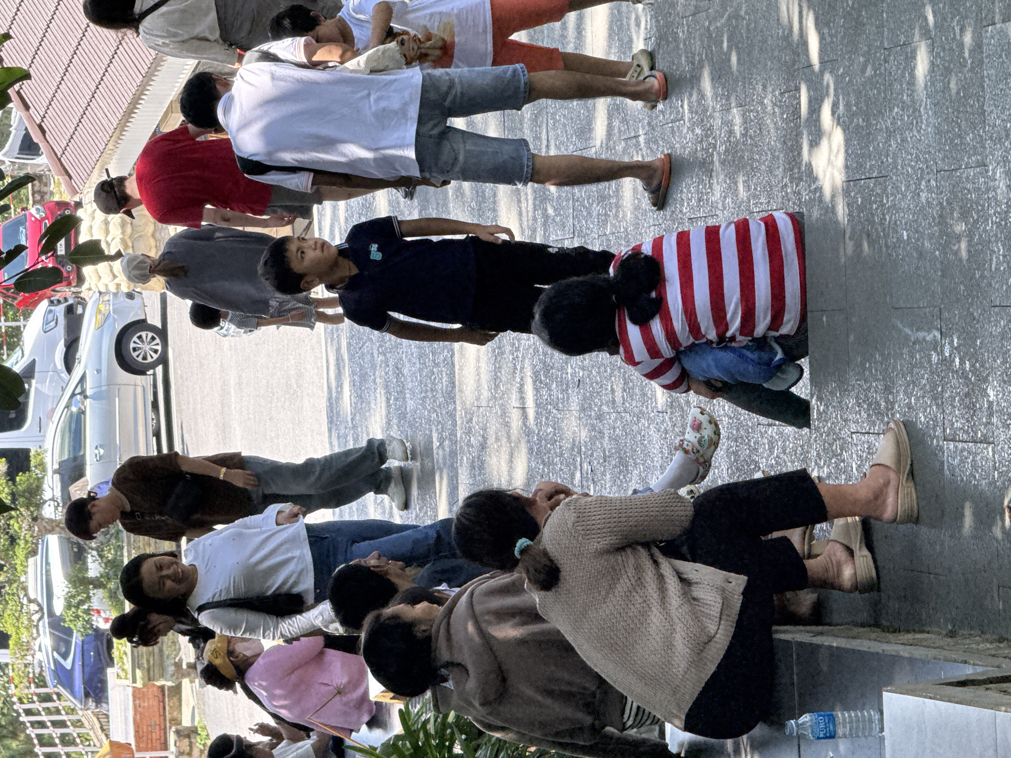 The image depicts a group of people gathered in a public outdoor space, likely in Da Lat, Vietnam, given the style of architecture and vegetation. The environment features a paved walkway with a textured surface, interspersed with patches of sunlight and shadow. In the background, a light-colored van and a red car are parked. Further back, a red-tiled roof structure is visible.
The people in the foreground are diverse in age and attire. Several women and children are present. One woman in the center foreground is wearing a white long-sleeved shirt and blue jeans, looking towards the viewer. To her left, another woman in a pink top and yellow hat is partially visible, sitting. To the right of the woman in white, a young boy in a navy blue polo shirt and black pants is looking to his left. Behind him, a man in a red t-shirt and shorts is walking away from the camera. Near the right edge of the image, a child in a white t-shirt and orange shorts is standing.
Other individuals are seated or partially obscured. Two women are sitting on a low wall or ledge to the left, one in a brown hooded sweatshirt and the other in a beige knitted sweater, both facing away from the camera. A child wearing a red and white striped shirt is seated on the ground in the lower right, facing away.
Objects in the scene include a clear plastic water bottle with a blue label on the ledge where the seated women are. The overall impression is a casual gathering or a stop on a tour, with people interacting and observing their surroundings.