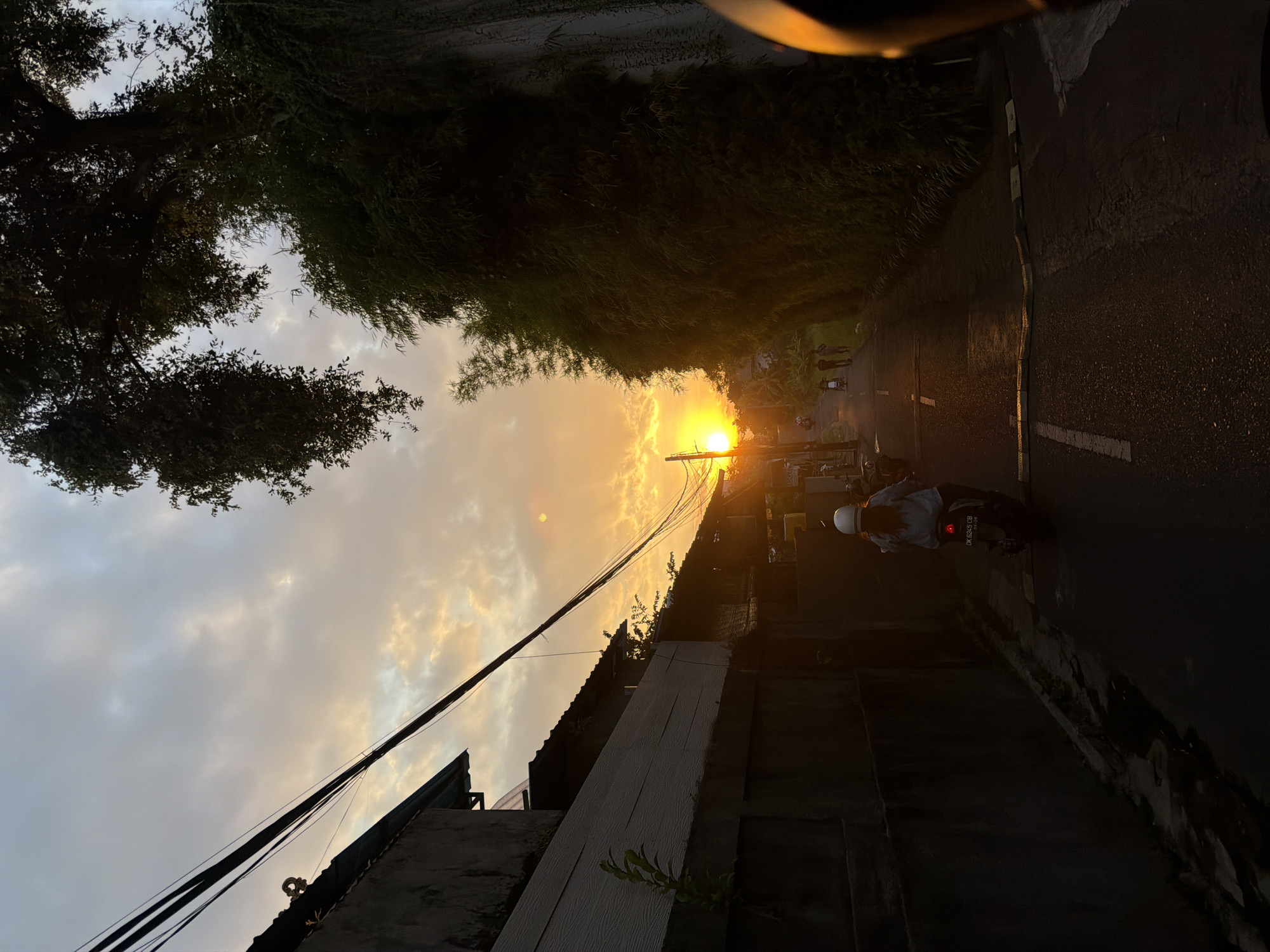 A motorcyclist wearing a white helmet rides down a narrow street during sunset in North Kuta, Indonesia. The sun, a bright orange orb, is visible in the sky, casting a warm glow on the clouds and illuminating the scene. The street is lined with buildings on the left and lush vegetation on the right, with power lines stretching across the sky. A few other people can be seen walking or riding motorbikes in the distance. The overall atmosphere is peaceful and serene, with a sense of everyday life unfolding as the day comes to a close.
