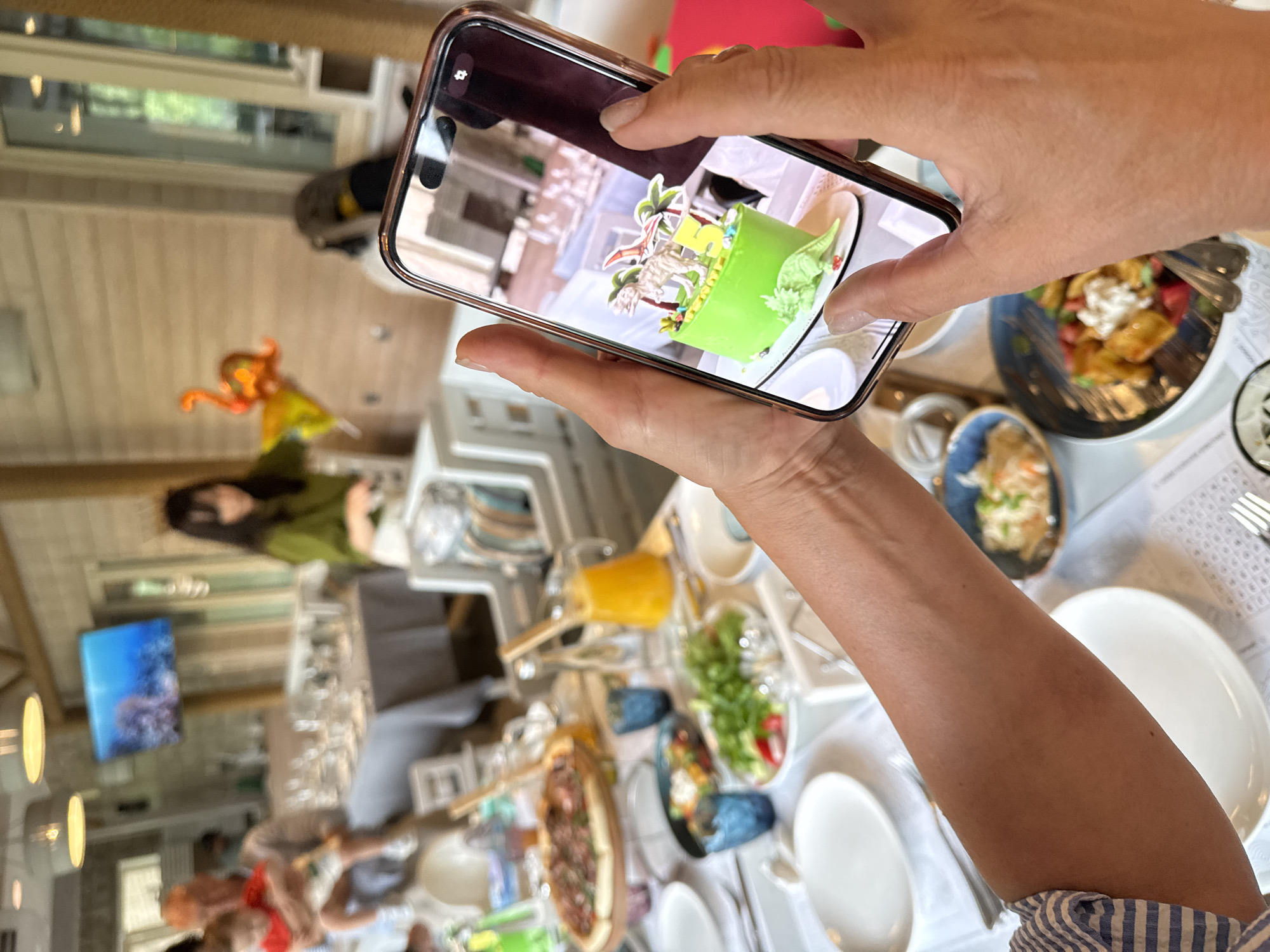 In this image, a person is holding a smartphone and taking a picture of a cake with a dinosaur theme. The cake is green and decorated with plastic dinosaur figures and palm trees, with a yellow number "5" prominently displayed. The scene takes place at a table laden with food, including a pizza, salads, and drinks in pitchers and glasses. In the background, other people are visible, including a man holding a child and a woman wearing a green shirt. A television screen displaying an underwater scene is mounted on the wall. The overall environment appears to be a restaurant or a large gathering space with wooden beams and paneled walls. The location is specified as Sovkhoza imeni Lenina, Russia.