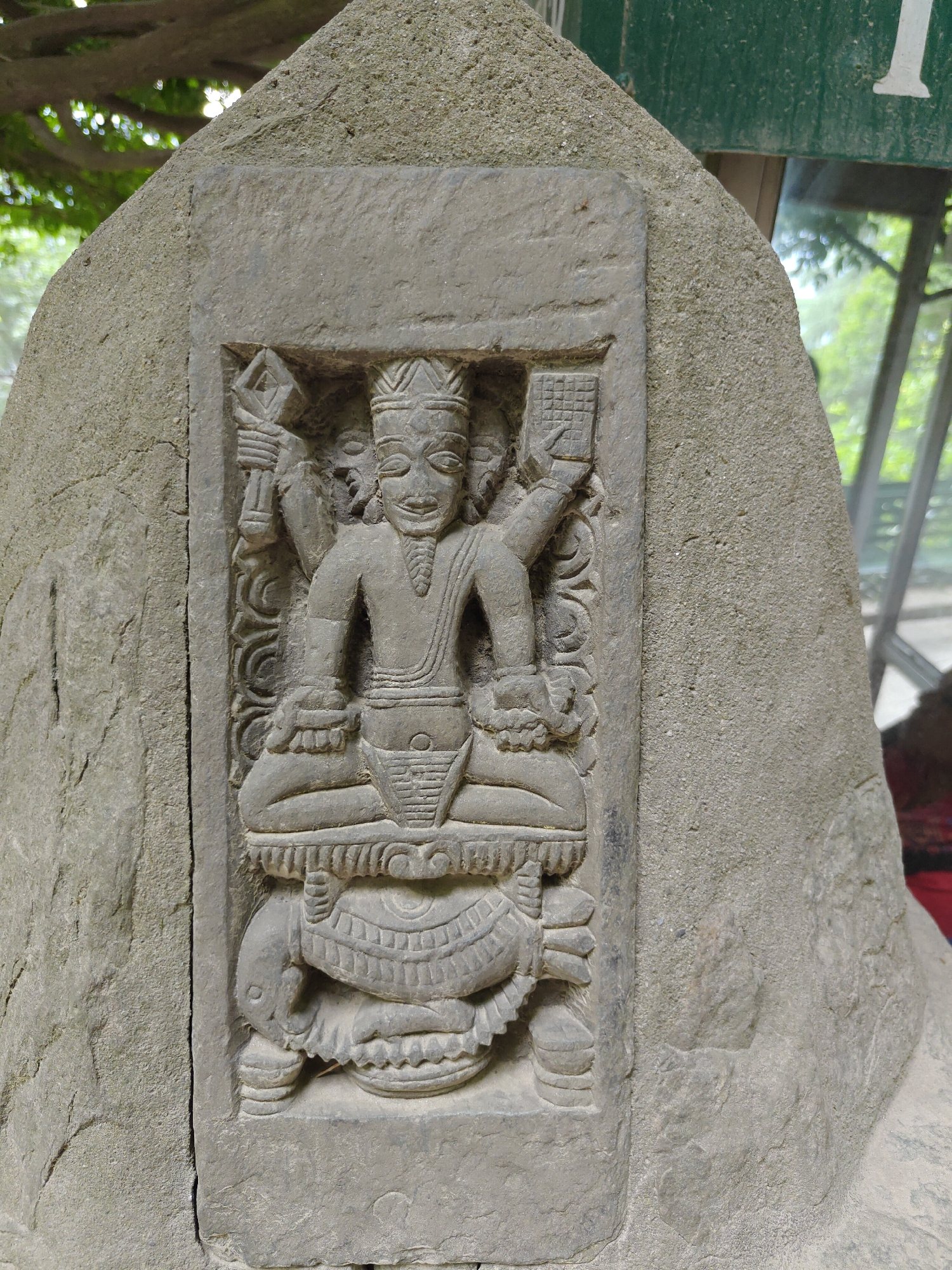 This image depicts a stone carving, likely part of a larger structure or monument, located in Naggar, India. The carving is of a seated figure with four arms. The figure appears to be male. The figure wears a crown, jewelry and is seated in a meditative pose atop an animal, possibly a tortoise or turtle. The background surrounding the carving is a natural stone structure. Parts of trees and a building are visible in the background suggesting an outdoor setting.