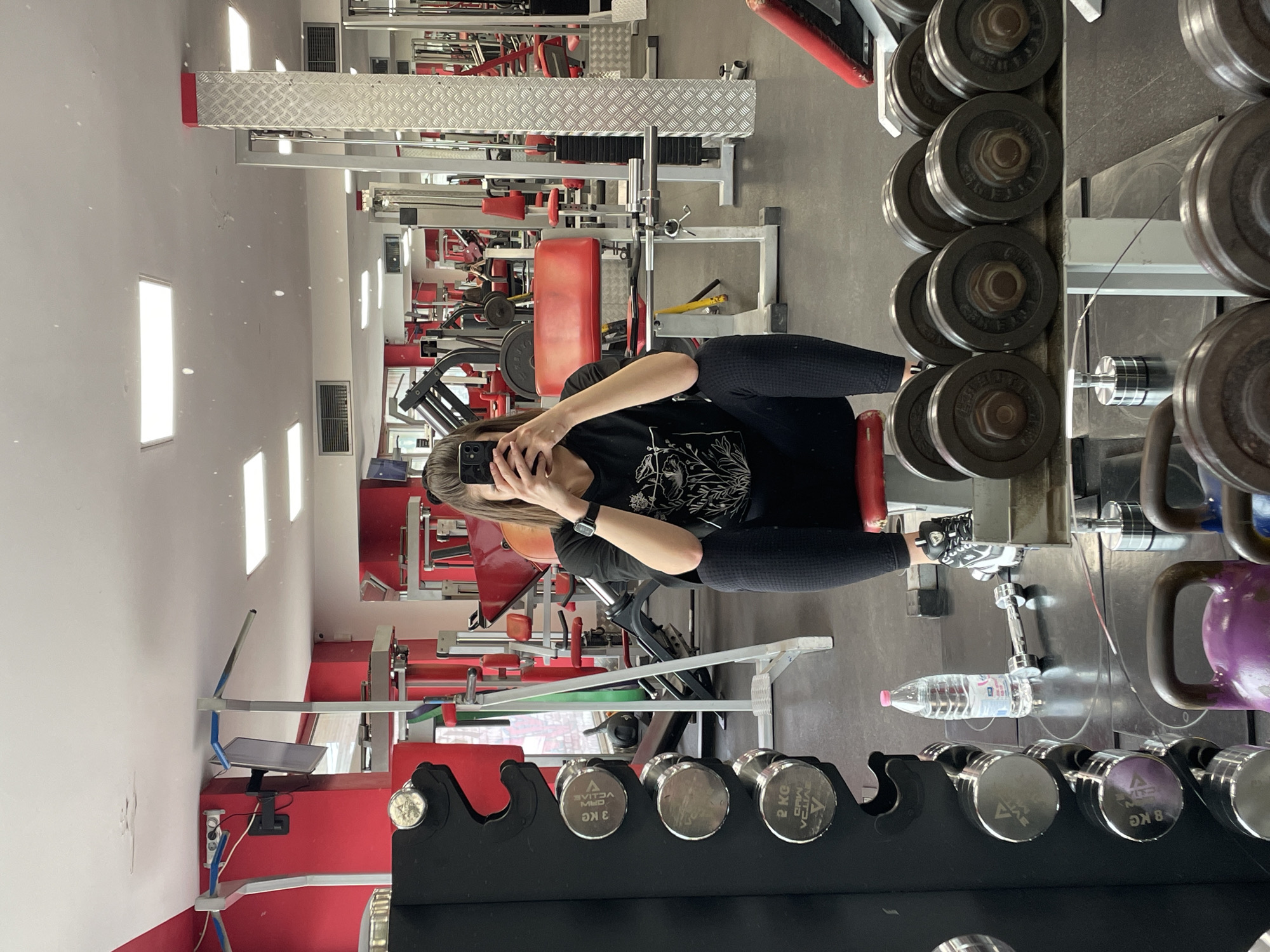 The image depicts a person taking a mirror selfie in a gym, likely located in Sofia, Bulgaria given the Cyrillic text on the weights. The gym has a variety of weightlifting equipment, including dumbbells, weight plates, and various machines. The walls are painted white with a red lower section. The person is wearing black leggings and a black t-shirt, holding their phone to take the photo.