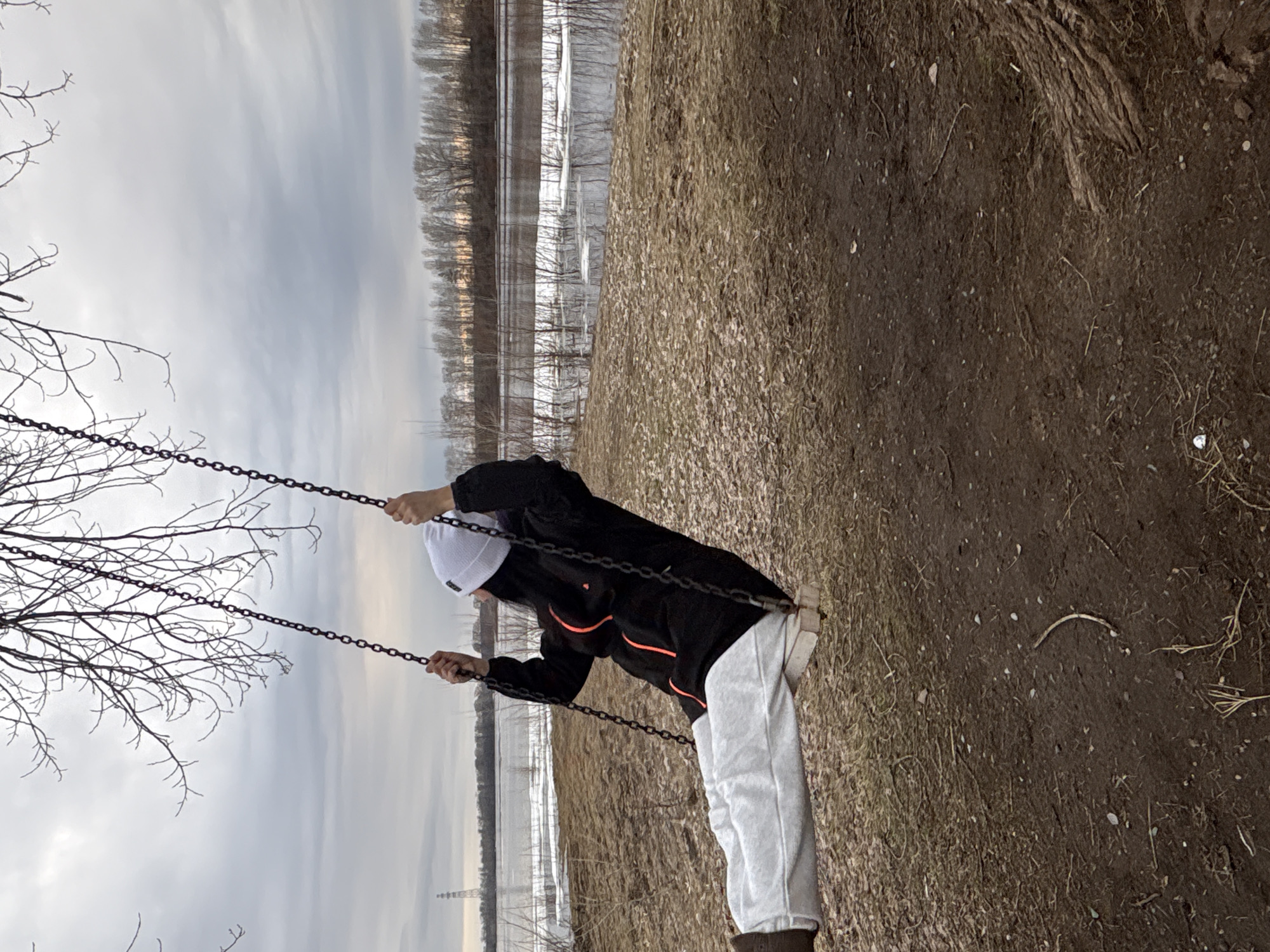 A person in a black jacket, light-colored pants, and a white cap is on a swing. The swing is located outdoors, possibly in a park. The chains of the swing are visible, and the person is holding onto them. The background includes trees, a body of water with some ice or snow, and an overcast sky. The ground appears to be a mixture of dirt and dead grass. The location could be Chaikovsky, Russia.